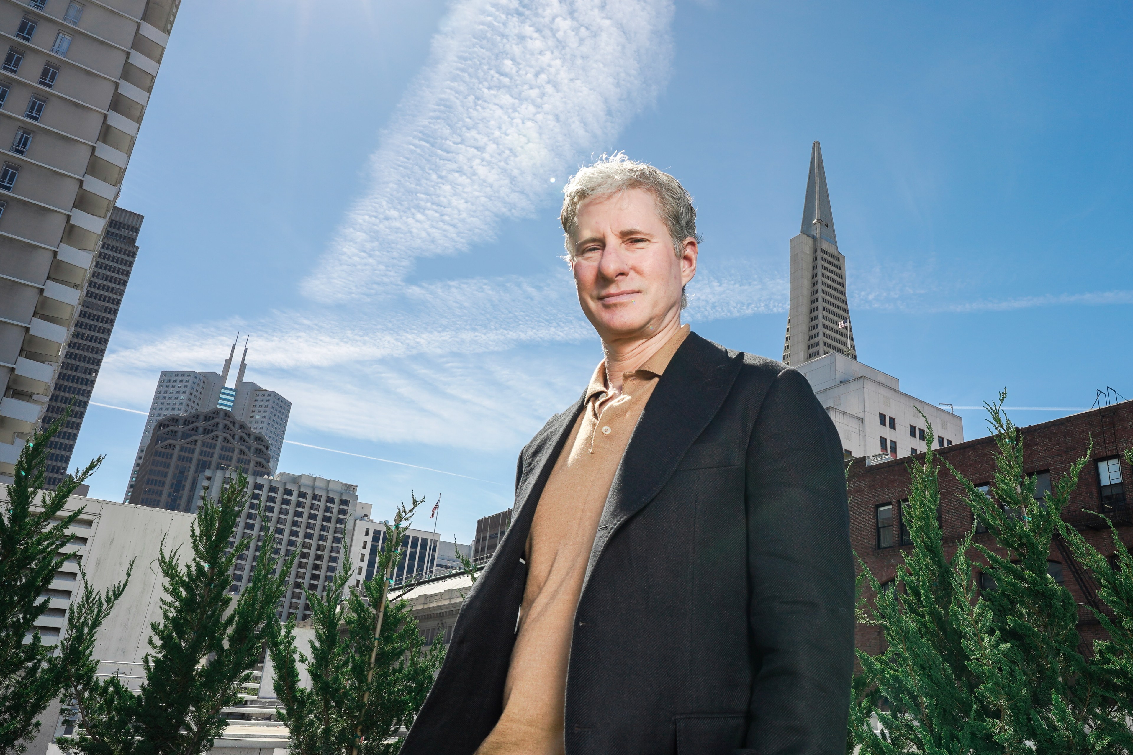 Chris Larsen stands before a cityscape with skyscrapers, a clear sky above, and greenery in the foreground.