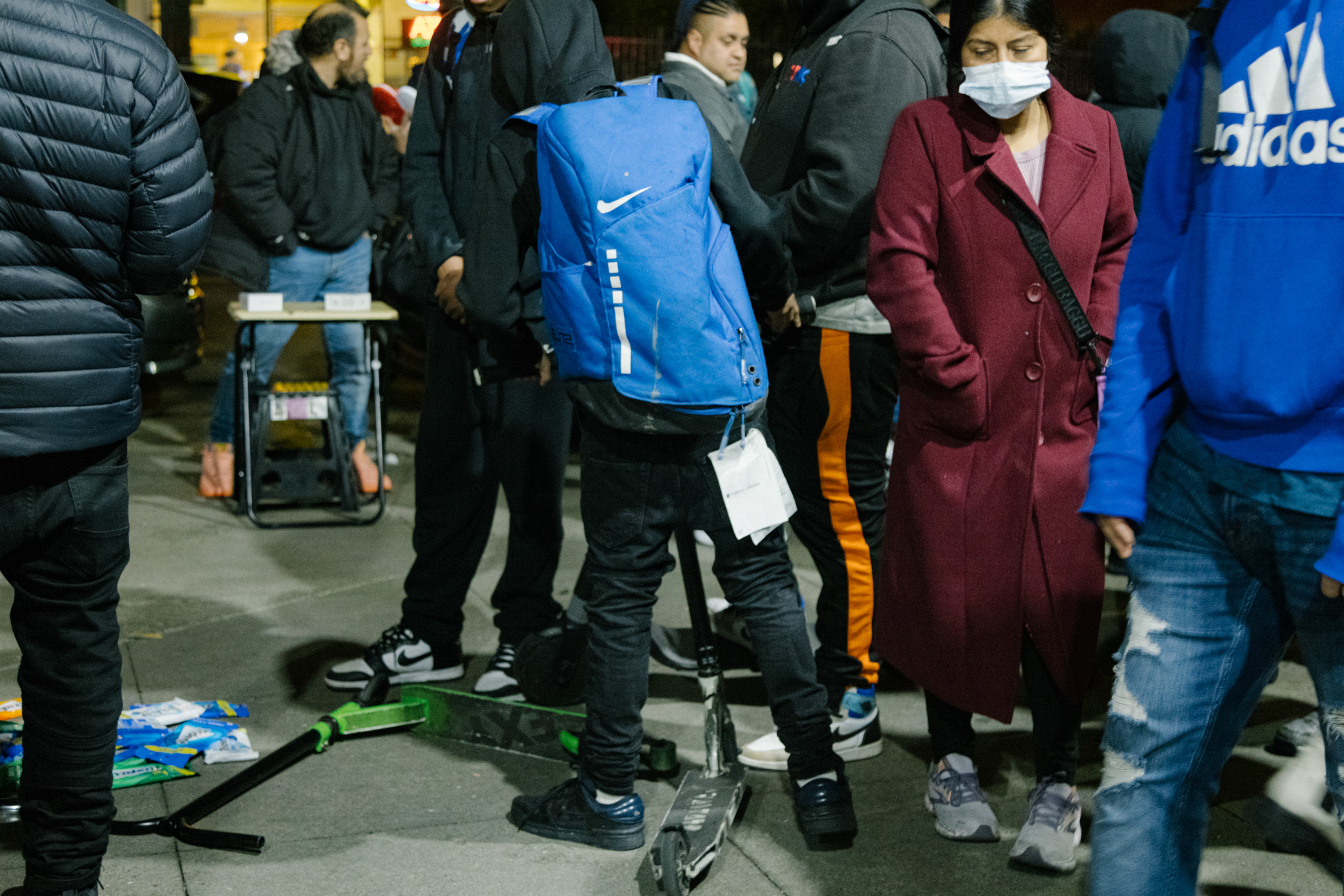 Crowded street scene at night with people walking and standing, some with backpacks and one wearing a mask.