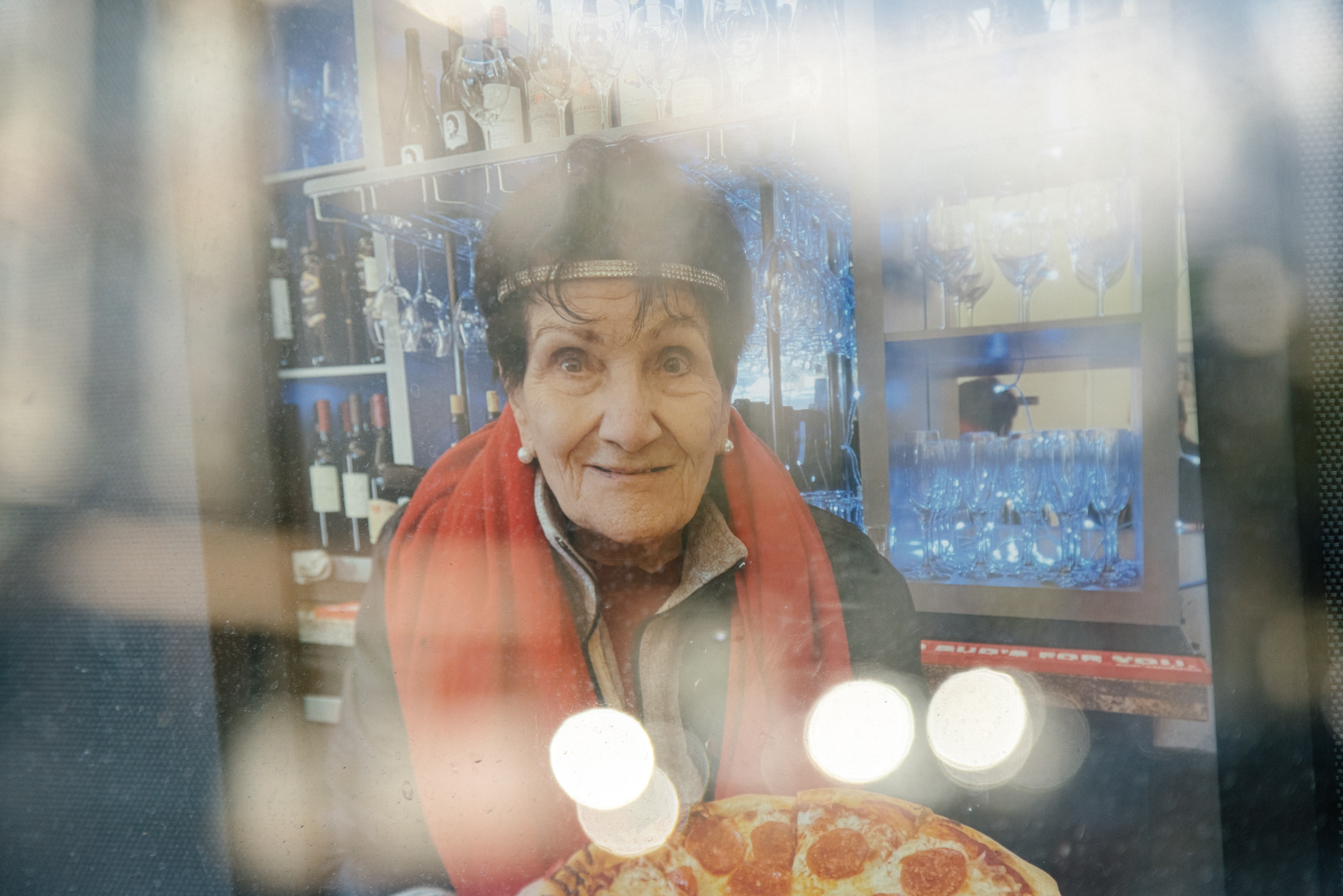 A smiling elderly person with a headband indoors, viewed through a glass window with reflections, beside a pizza on a table.
