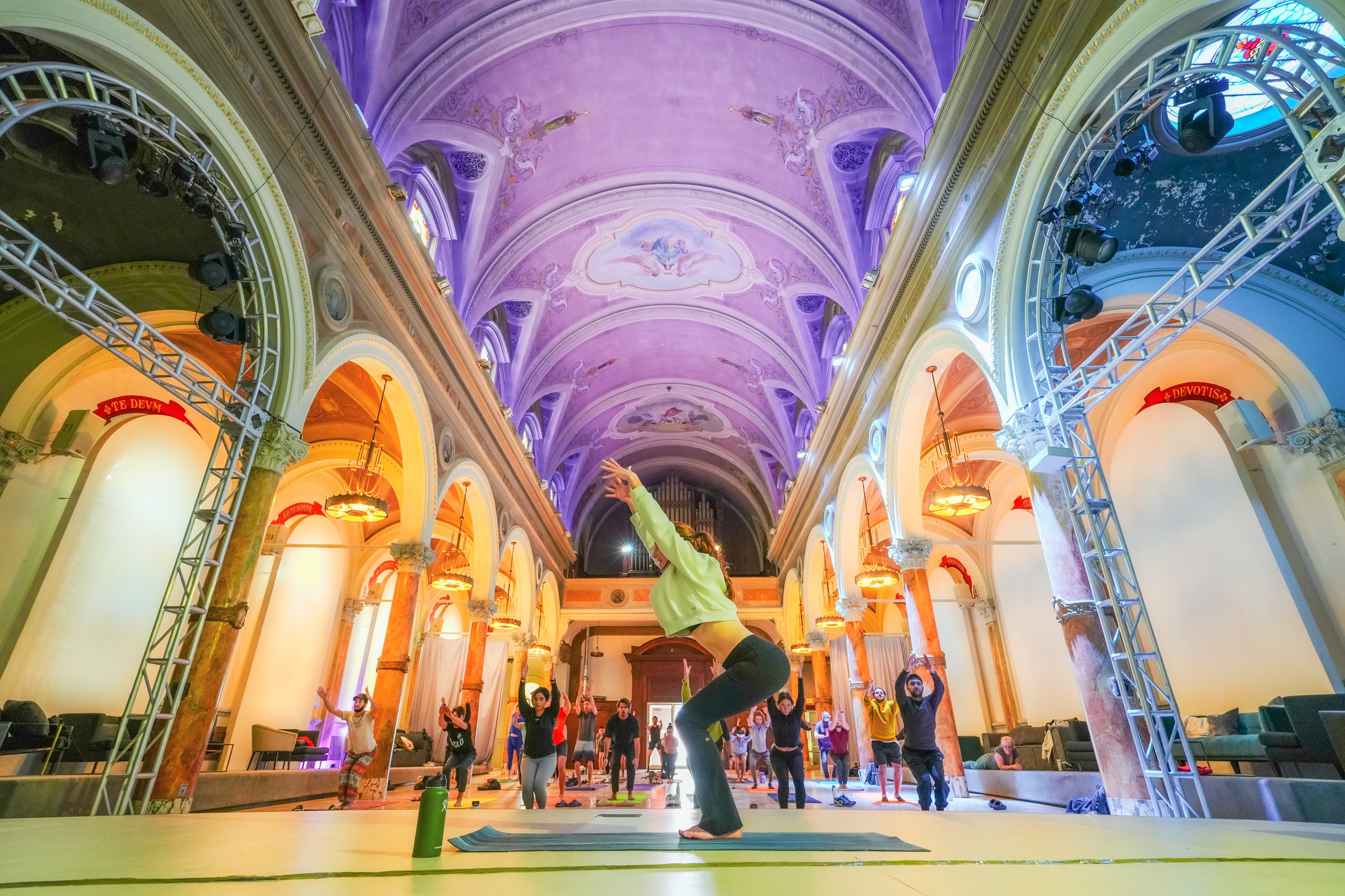 A yoga class inside a grand, ornate hall with violet ceilings, arches, and people in various poses.
