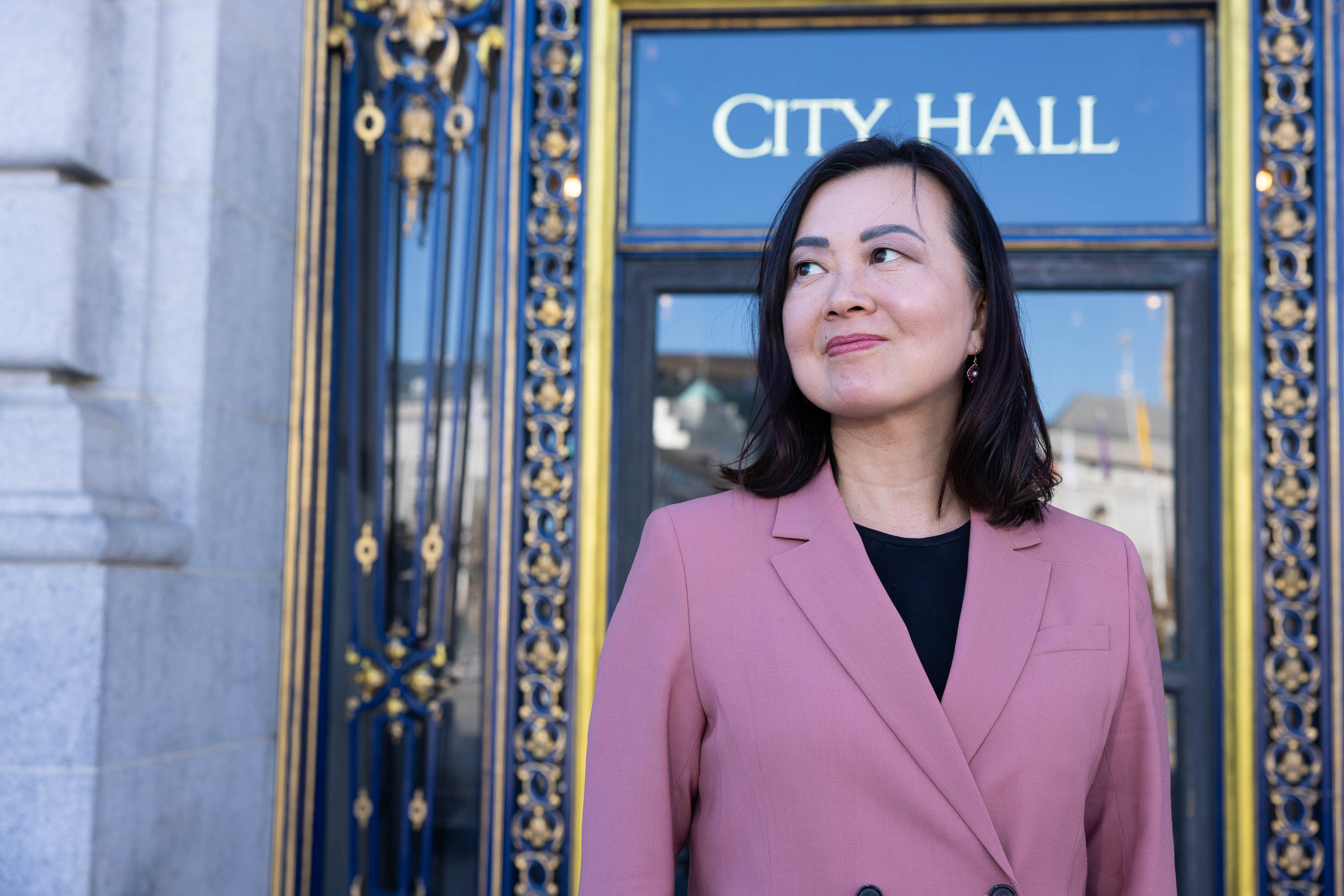 A woman in a pink blazer stands before a "City Hall" sign on a blue, gold-trimmed door.