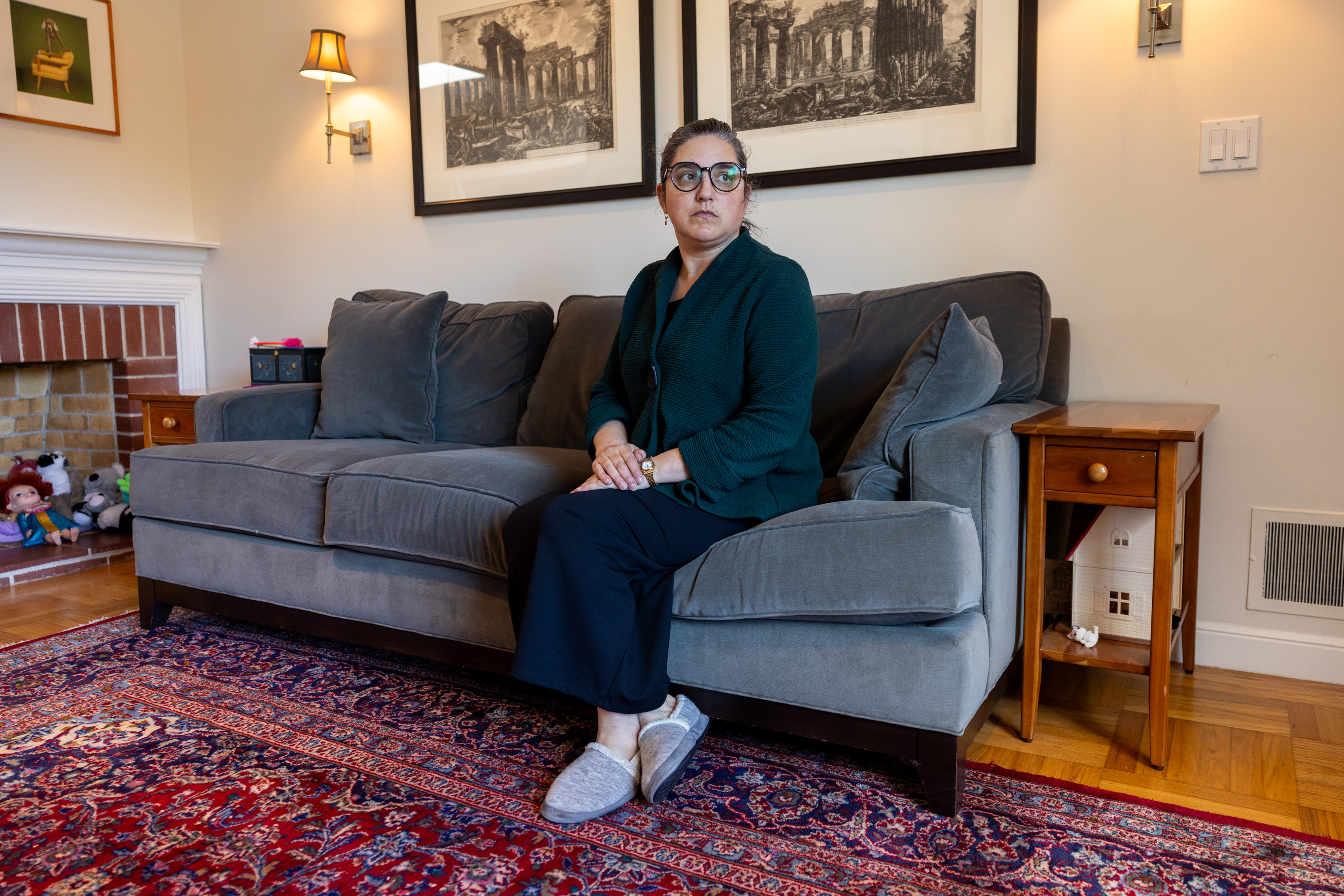 A woman sits on a gray couch in a cozy room with a Persian rug, artworks, and plush toys.