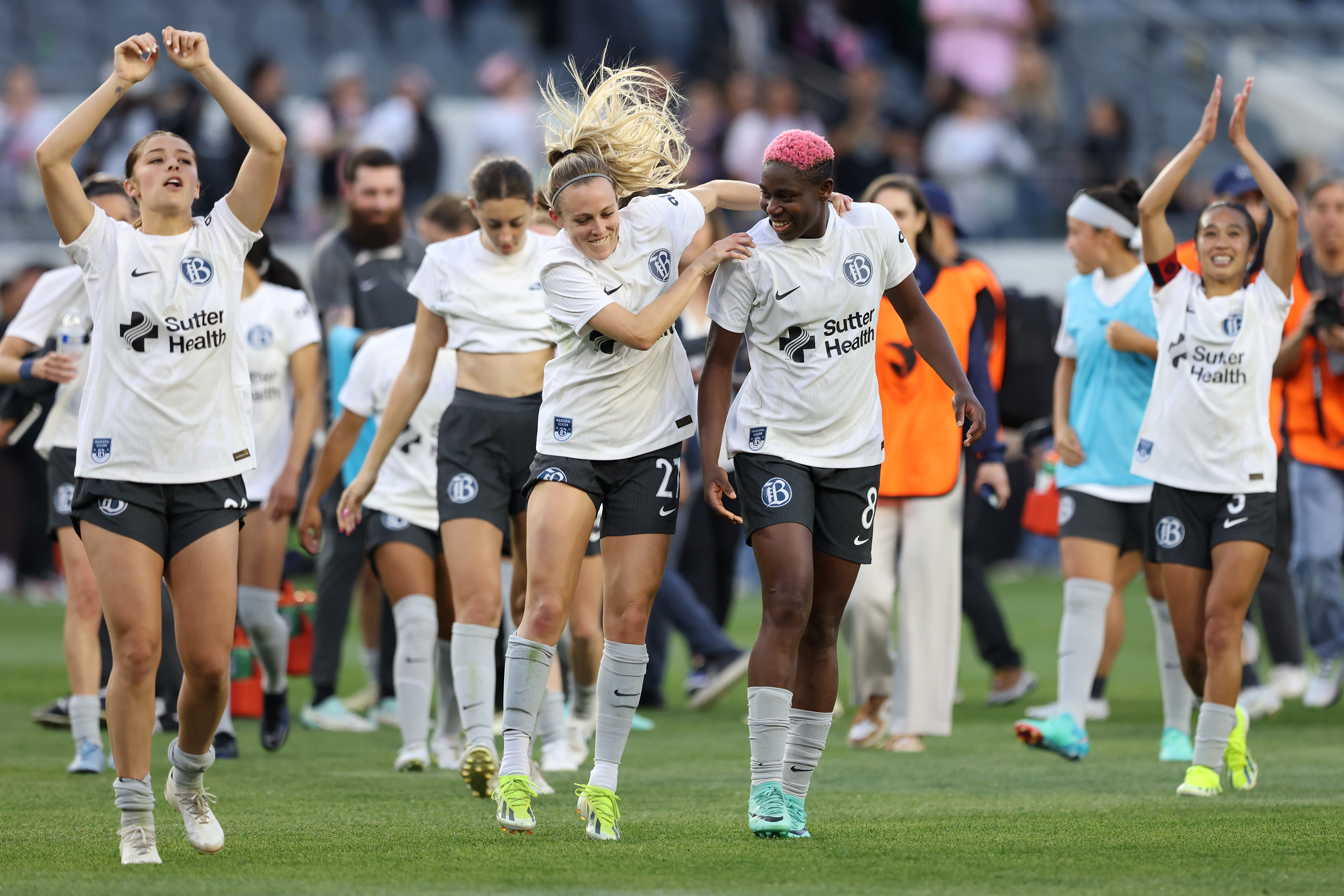 A group of female soccer players in white kits joyfully walk on a field, celebrating with raised arms and smiles.