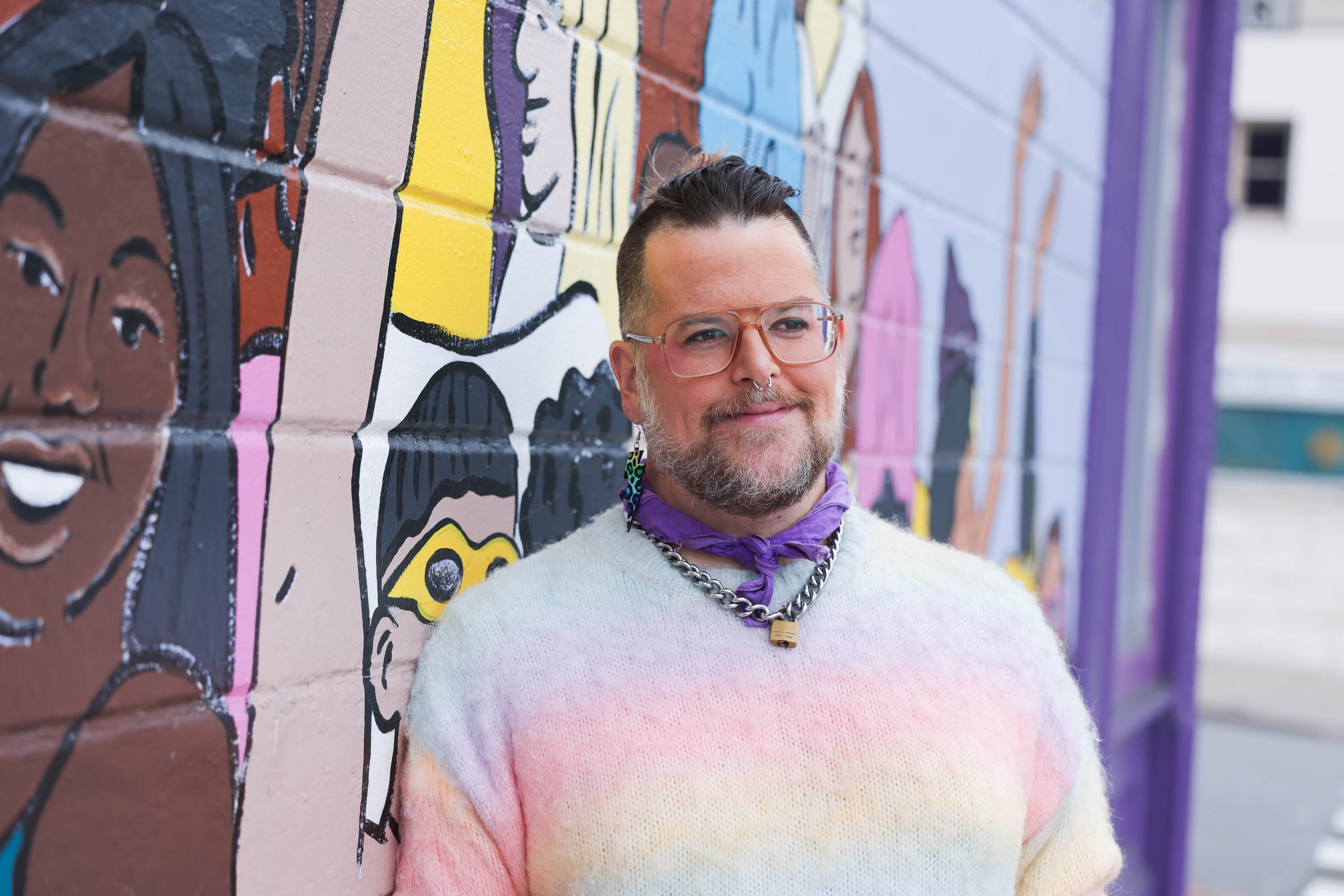 a smiling nonbinary person in glasses and a pastel sweater stands in front of a colorful exterior mural at an oblique angle.