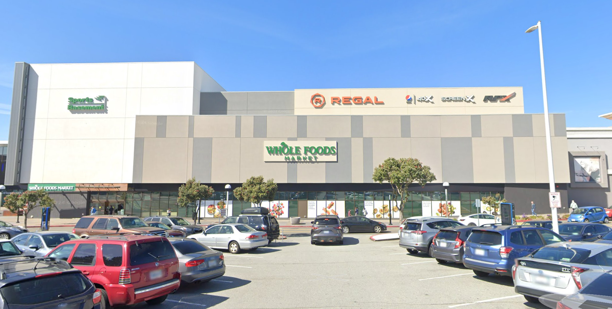 A parking lot in front of a mall with signs for Whole Foods and Regal Cinemas under a clear sky.