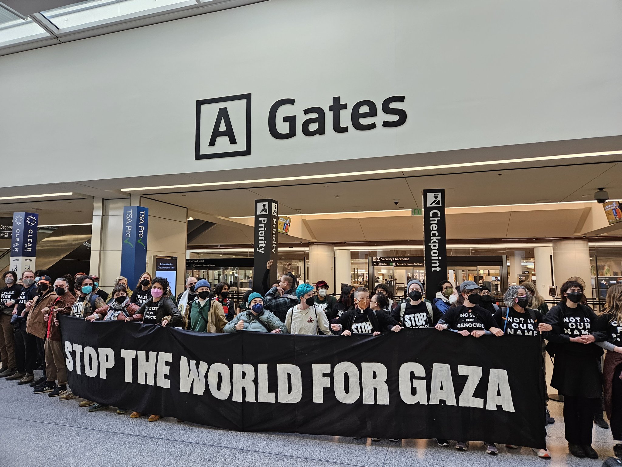 Group of masked people holding a banner reading "STOP THE WORLD FOR GAZA" at an airport gate area.