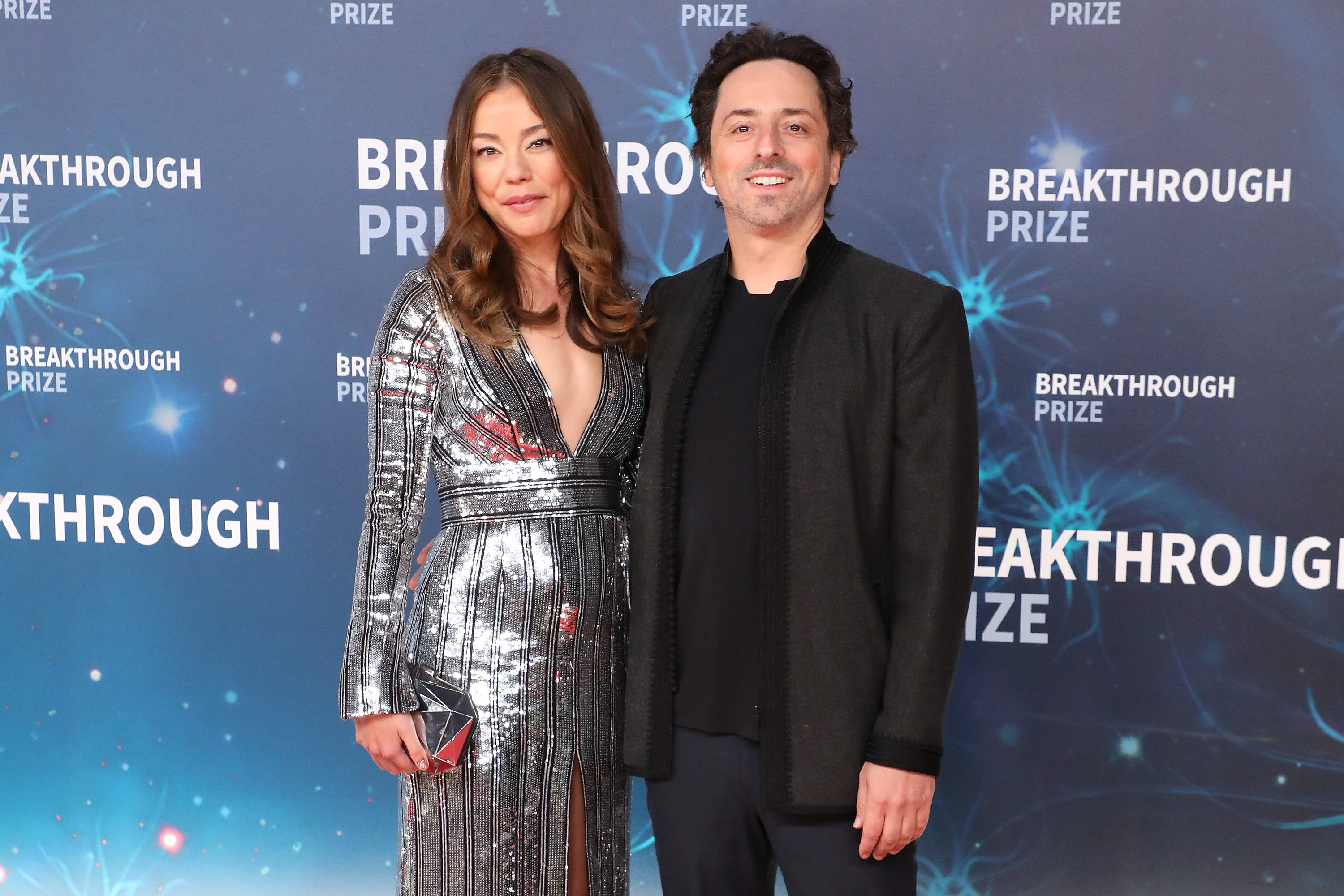 A man and woman stand together against a backdrop with &quot;Breakthrough Prize&quot; written, both dressed for a formal event.