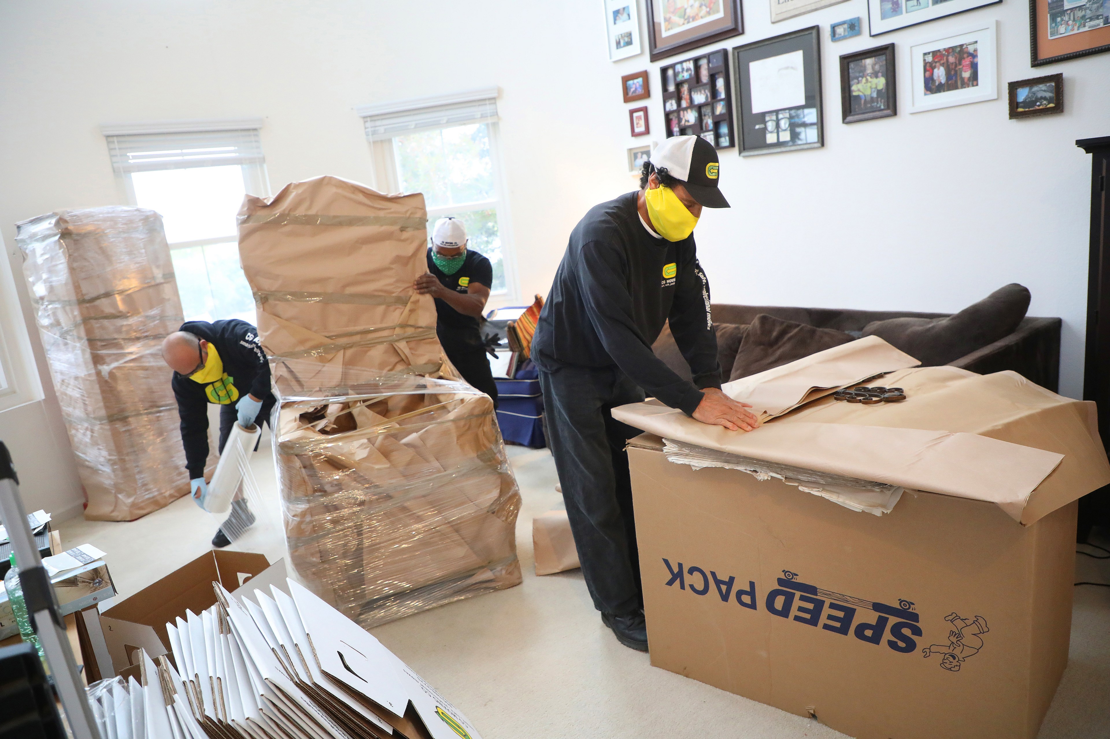Three movers are packing/unpacking items in a room with framed photos on the wall.