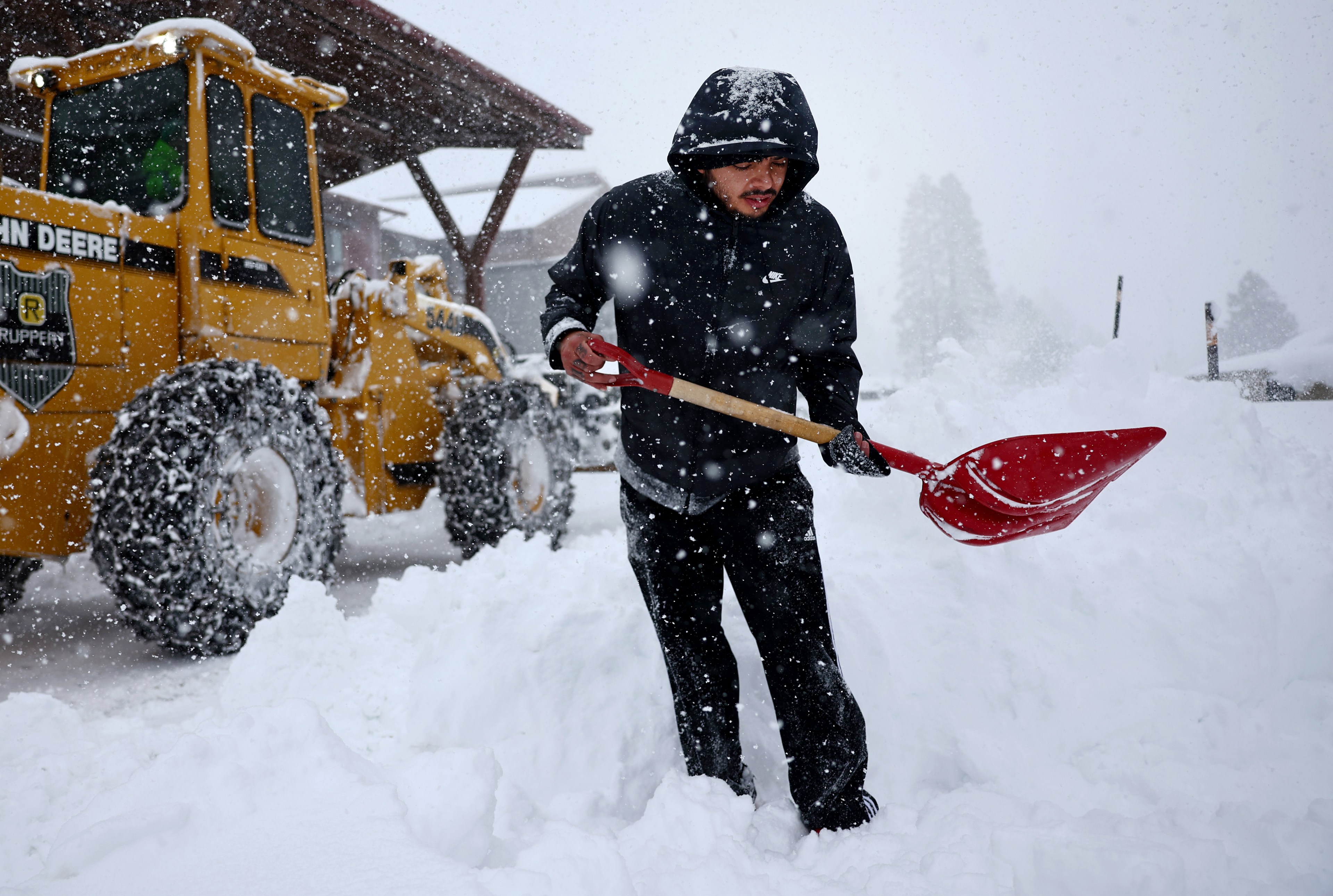 Man standing in knee deep snow shovels snow in front of yellow tractor