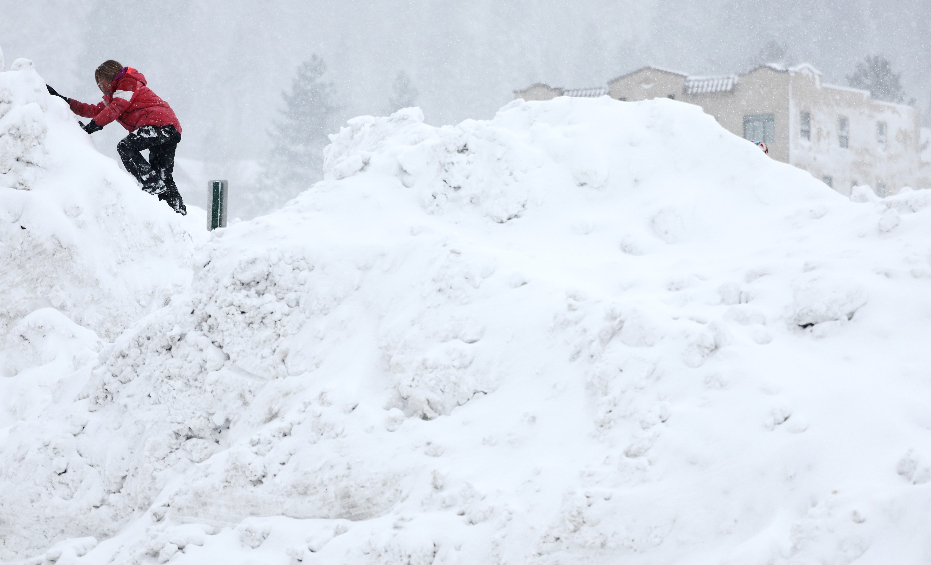 A child in a red jacket and black pants climbs on top of pile of snow