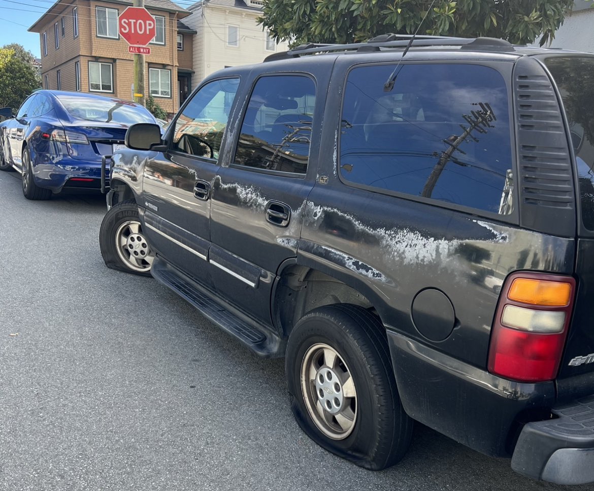 A black SUV with faded paint and scratches, parked on a street beside a blue car near a stop sign.