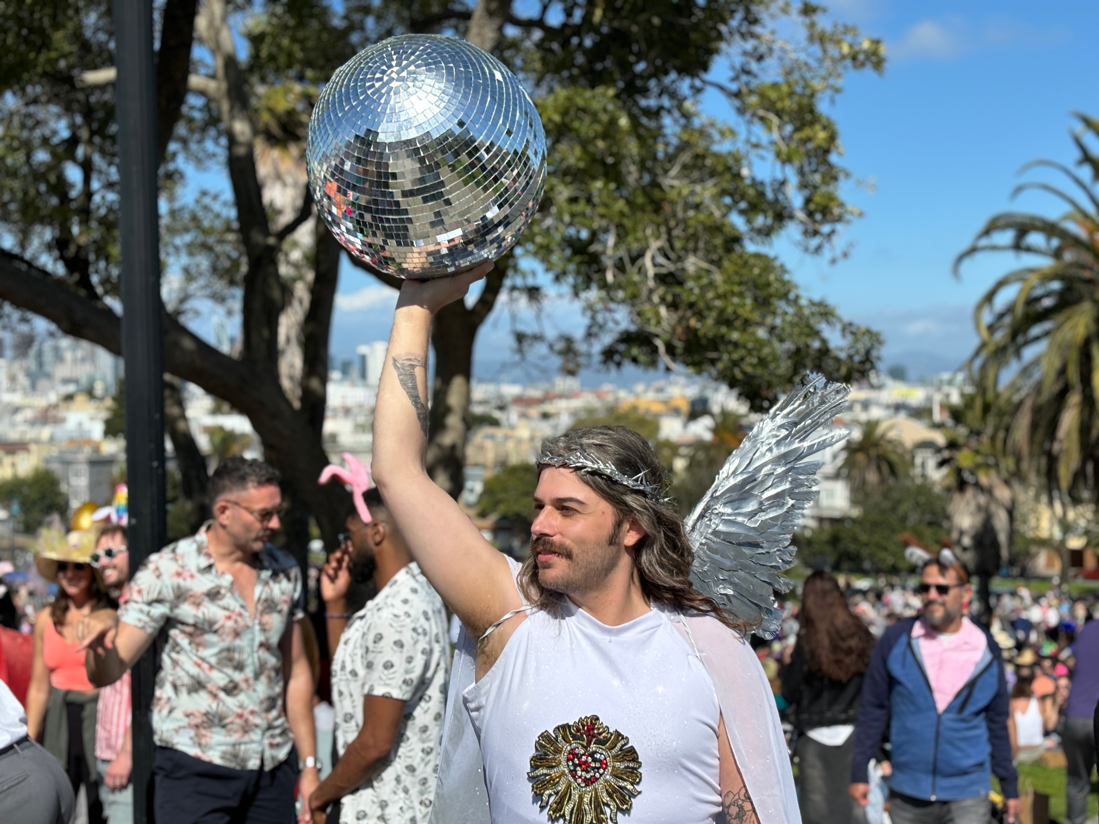 A person with a silver wing headband and wings raises a disco ball outdoors among a festive crowd.