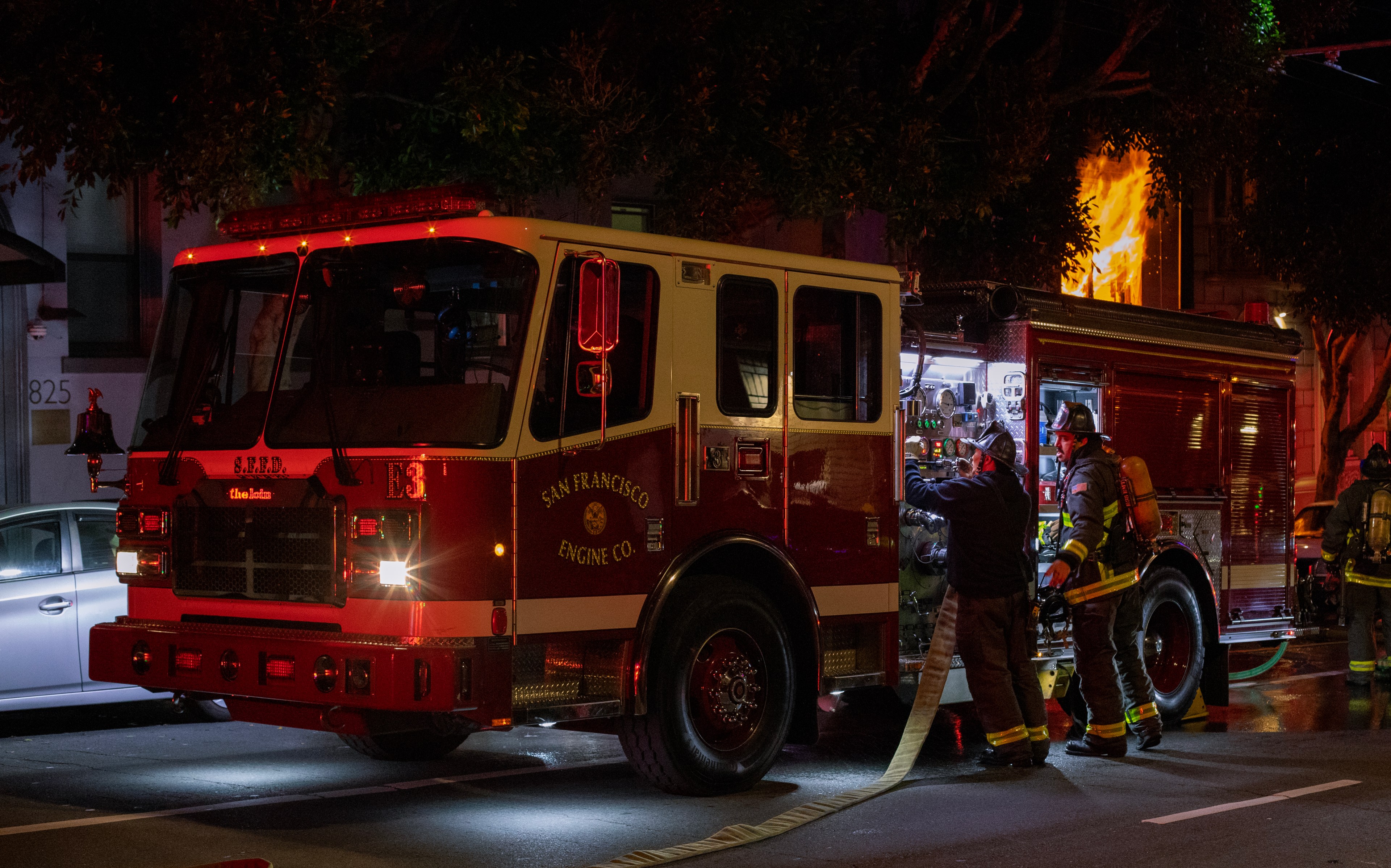 Firefighters stand by an engine at night parked outside an apartment building with fire coming from a window.