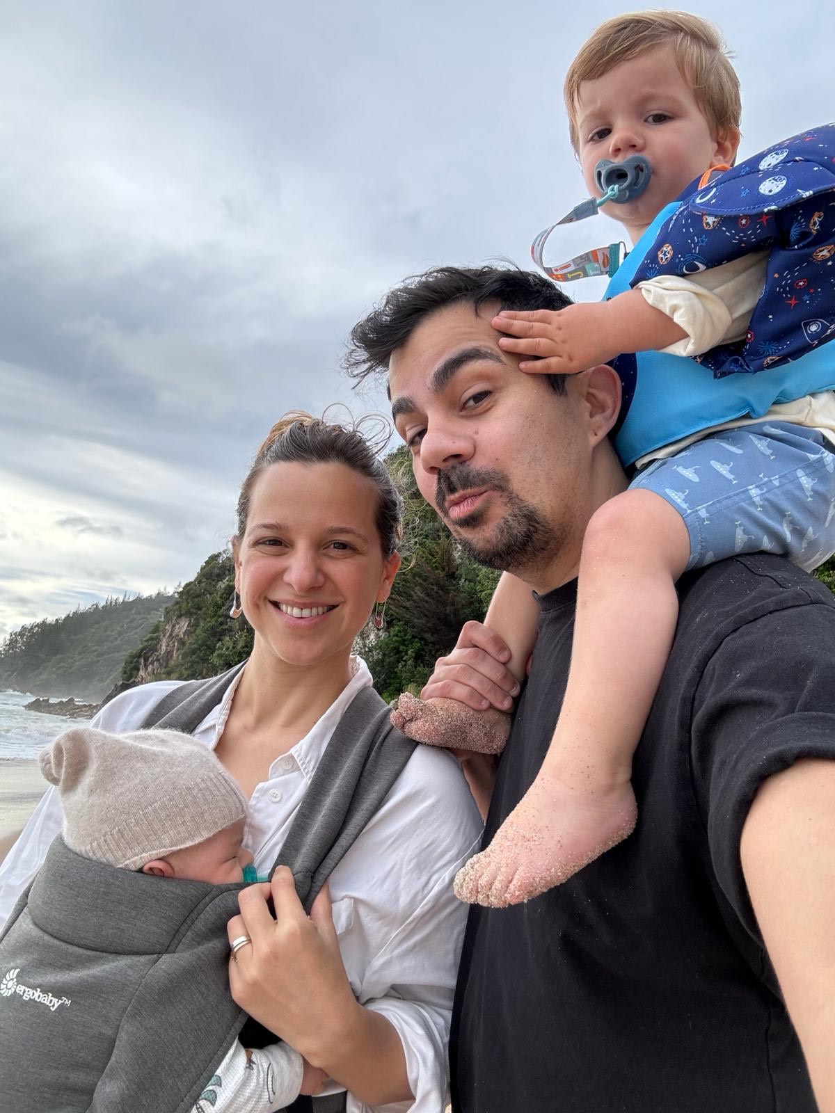 A family with two kids on a beach: one infant in a carrier and a toddler on his dad's shoulders.