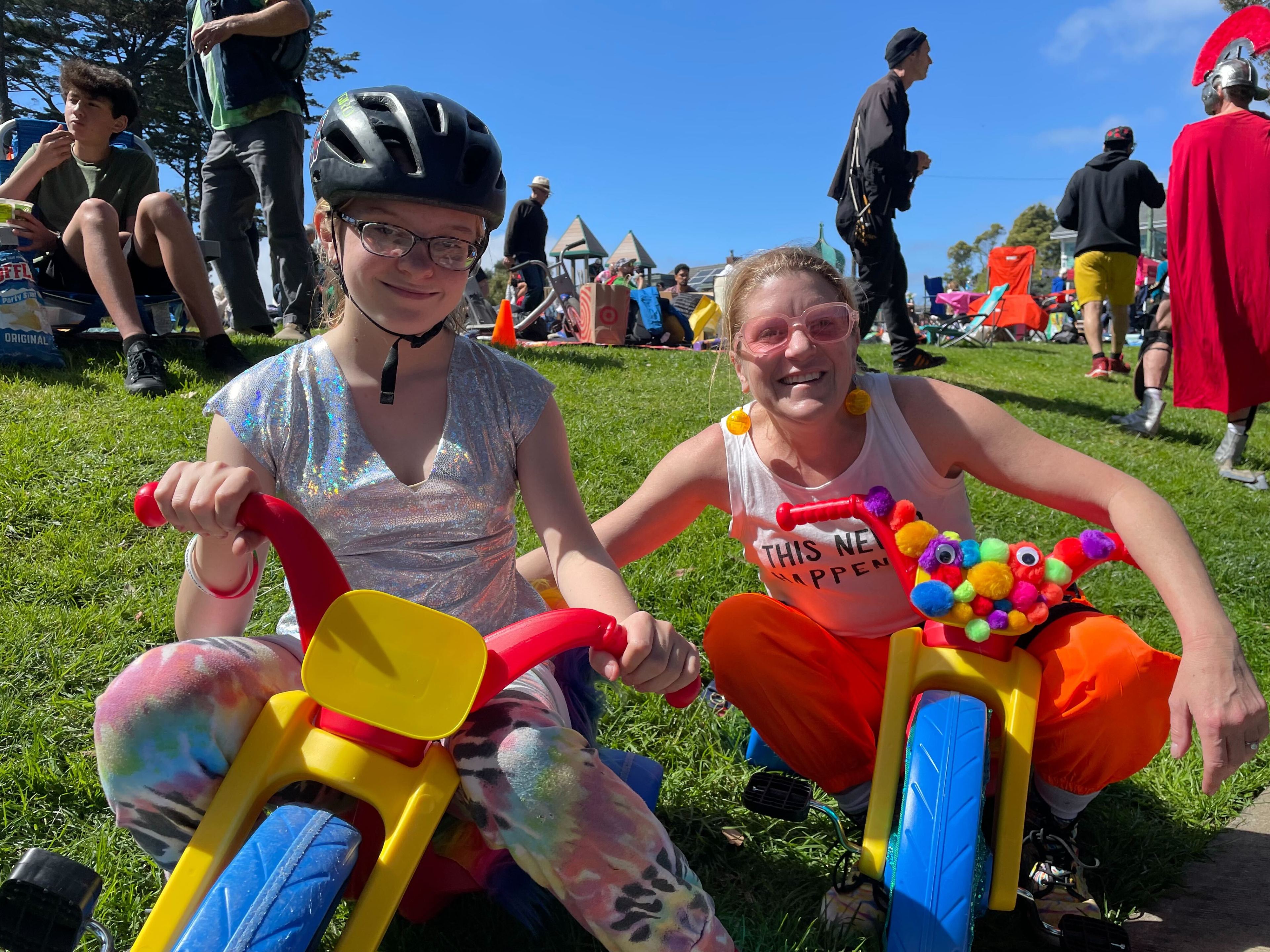 Two people are smiling on a sunny day, one on a small toy bike, both wearing colorful, quirky outfits and helmets.