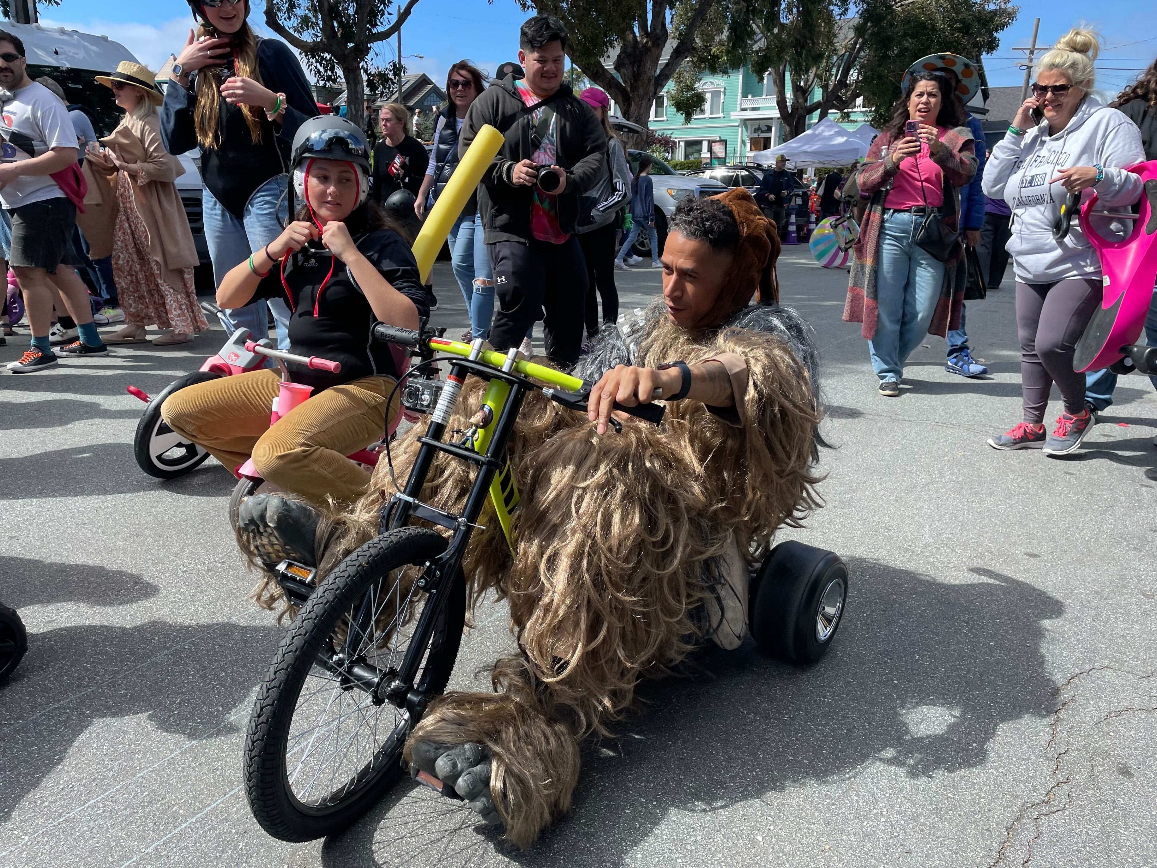 A person in a furry costume rides a tricycle with onlookers at a street event.