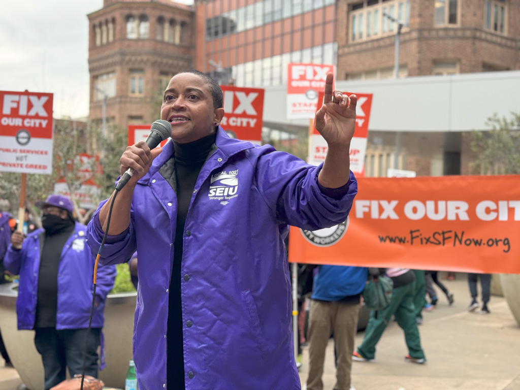A person in a purple jacket speaks into a mic at a rally with a "FIX OUR CITY" banner in the background.