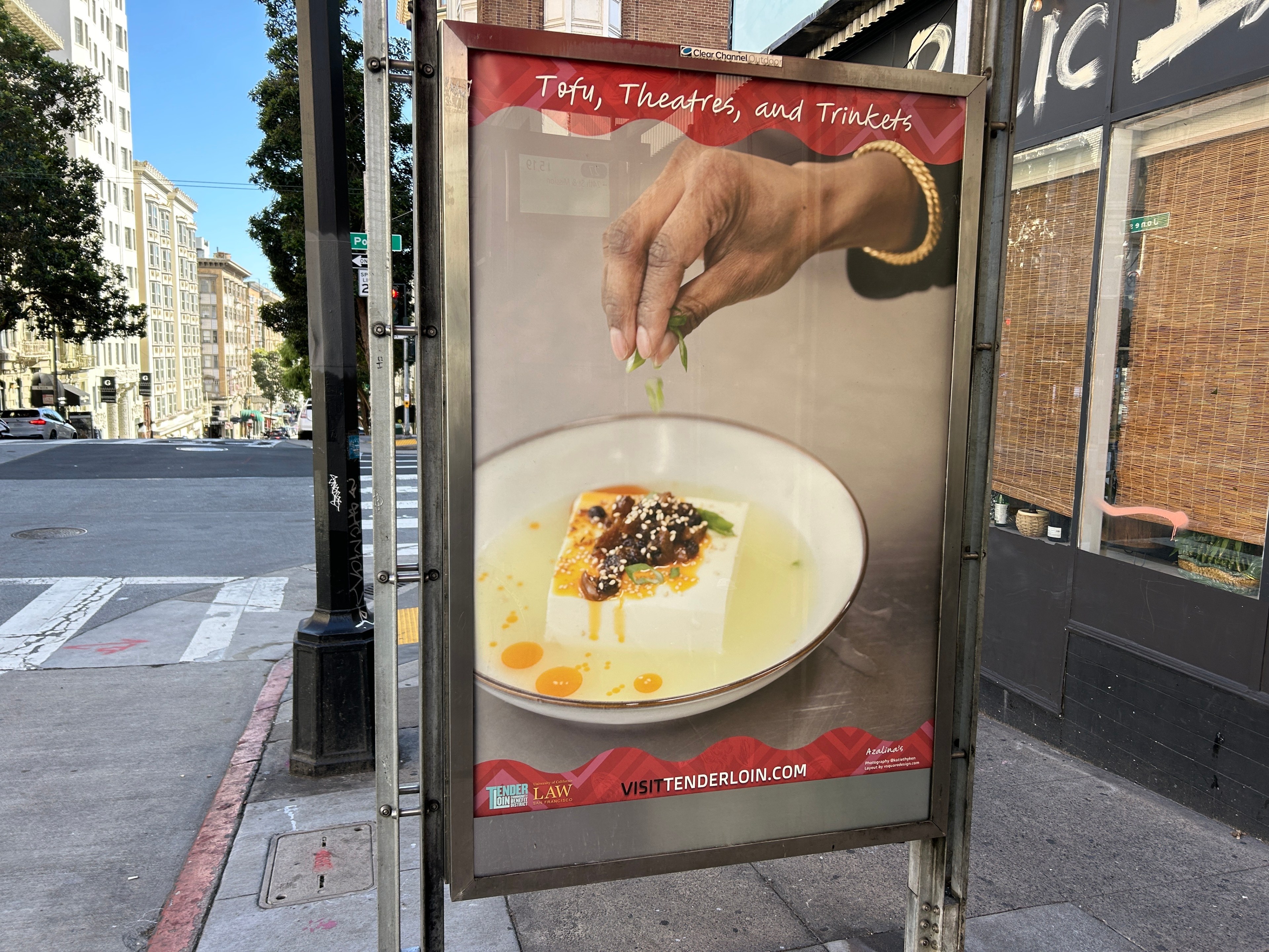 An outdoor ad depicts a hand garnishing a dish, with the text &quot;Tofu, Theatres, and Trinkets&quot; above. It's on a city street corner.