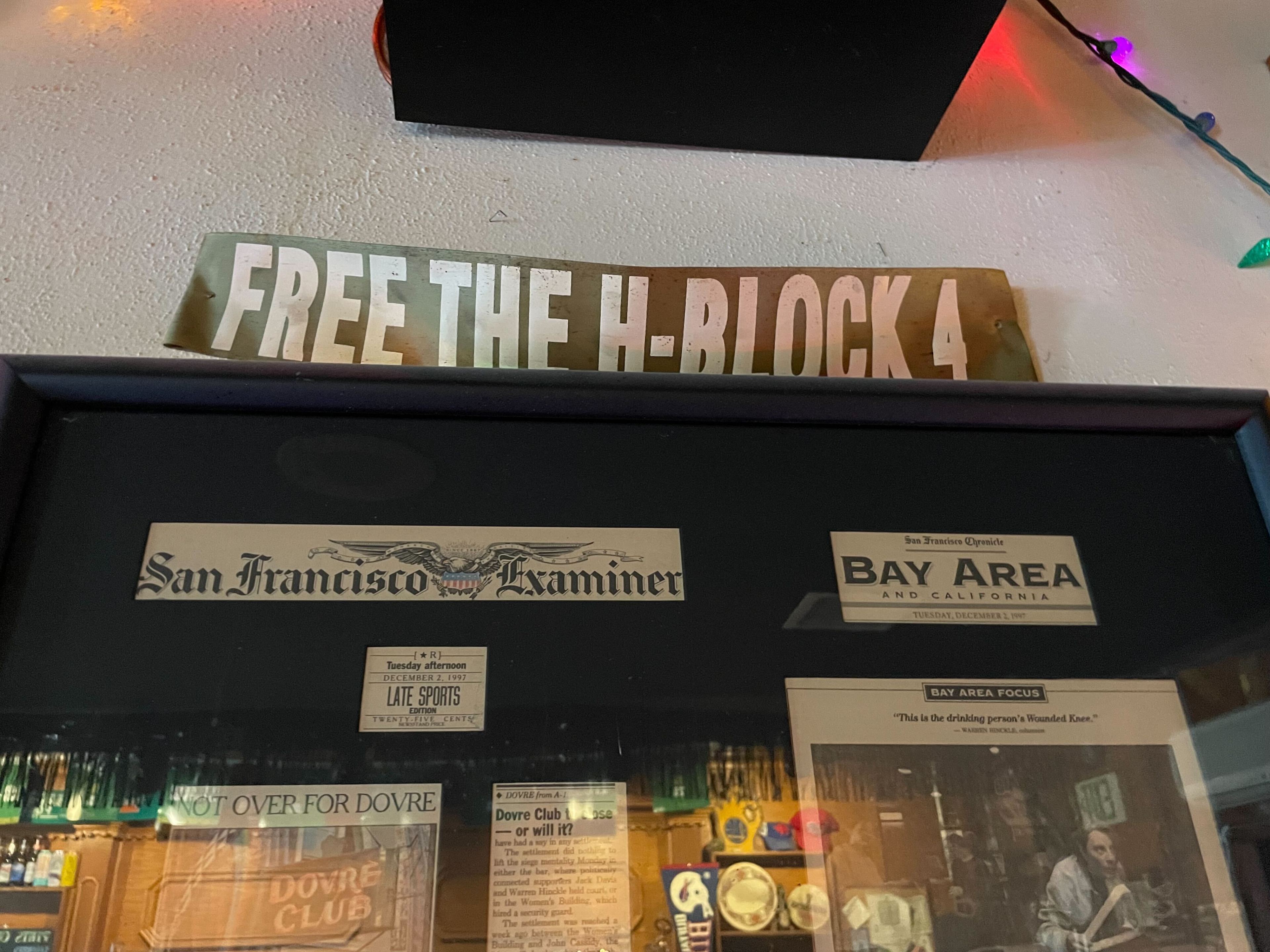 A sign saying &quot;FREE THE H-BLOCK 4,&quot; with framed newspaper titles below, and Christmas lights above.