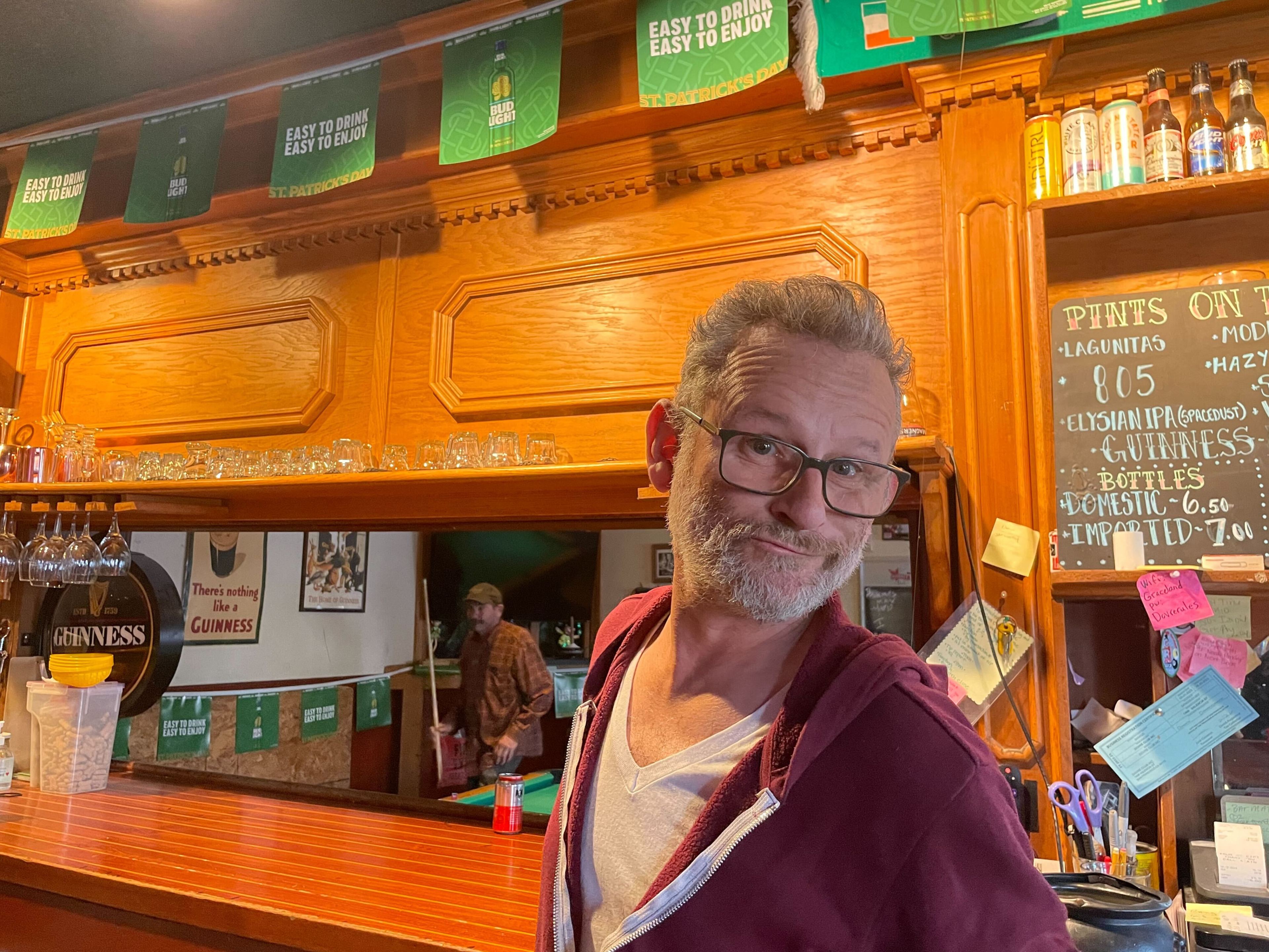 A man leaning on a pub bar with beer taps, glasses, and a price board in the background.