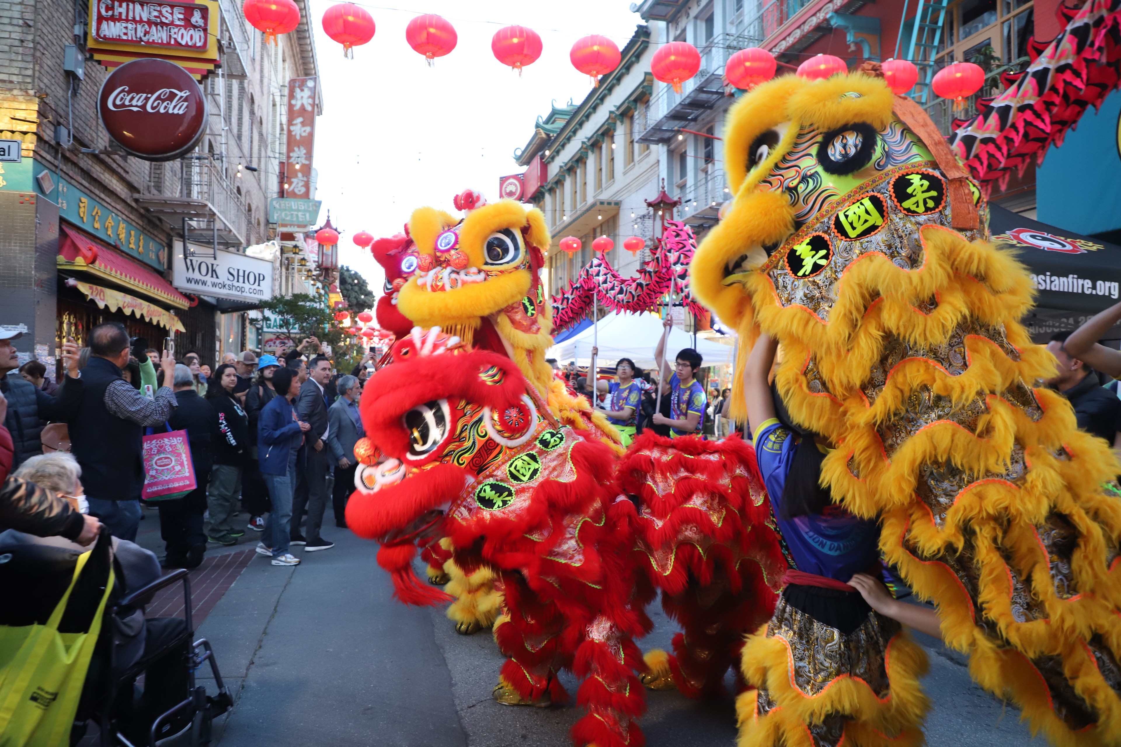 Two vibrant lion dance costumes perform in a bustling street with red lanterns and onlookers.