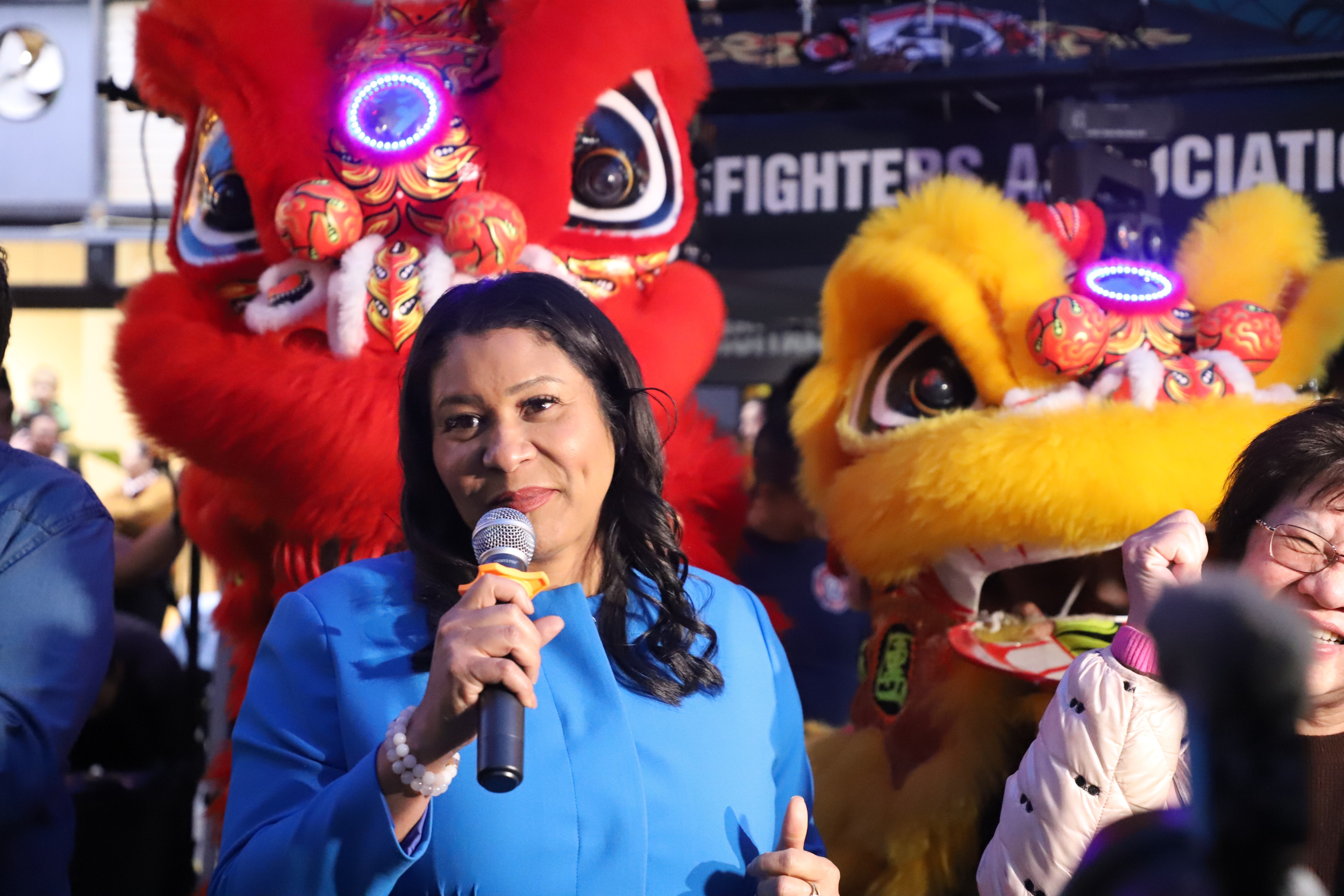 A woman speaks into a mic, smiling, with colorful lion dance costumes behind her.