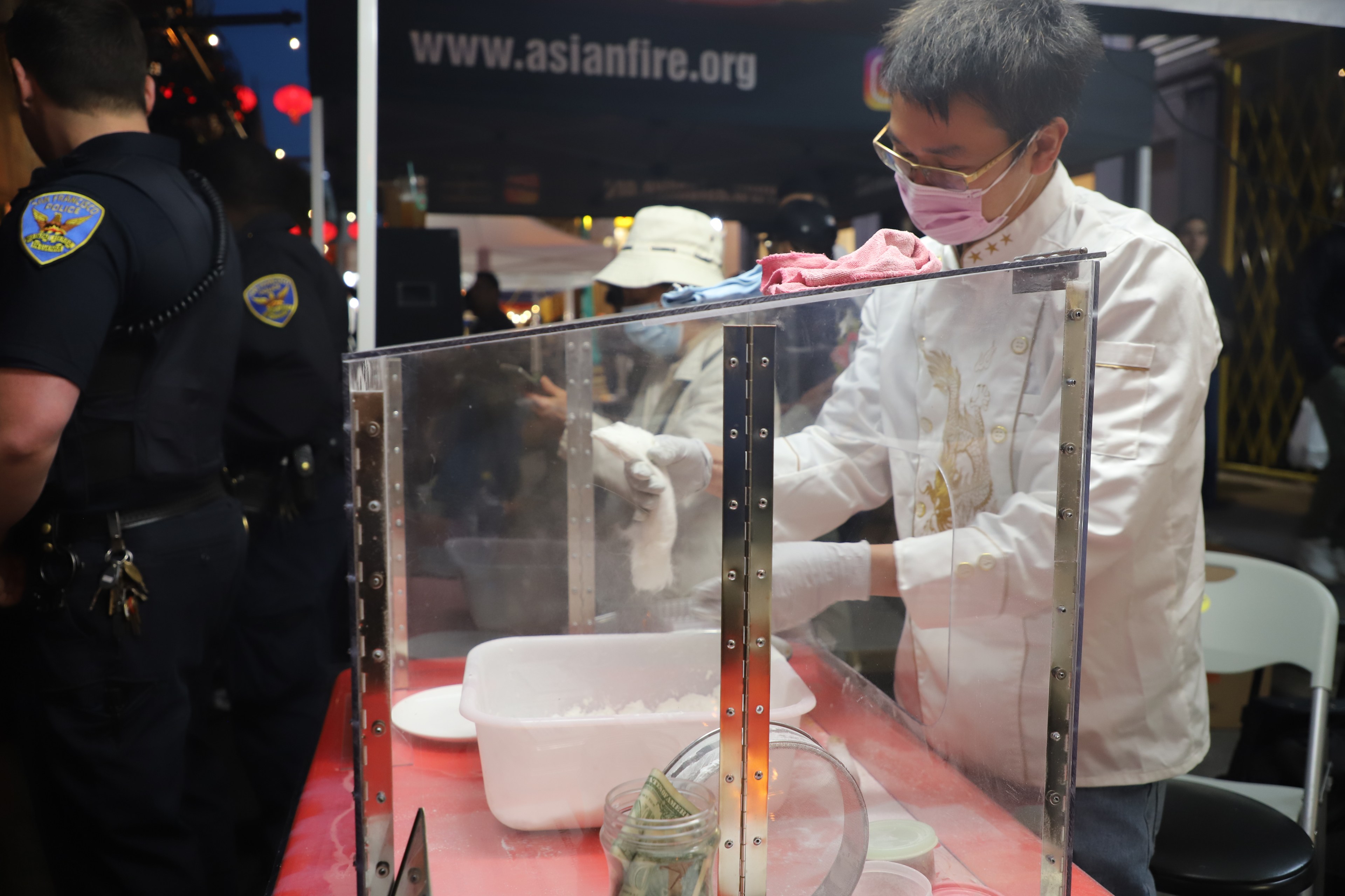 A person in a white chef's jacket and mask making food behind a clear barrier, watched by police officers.