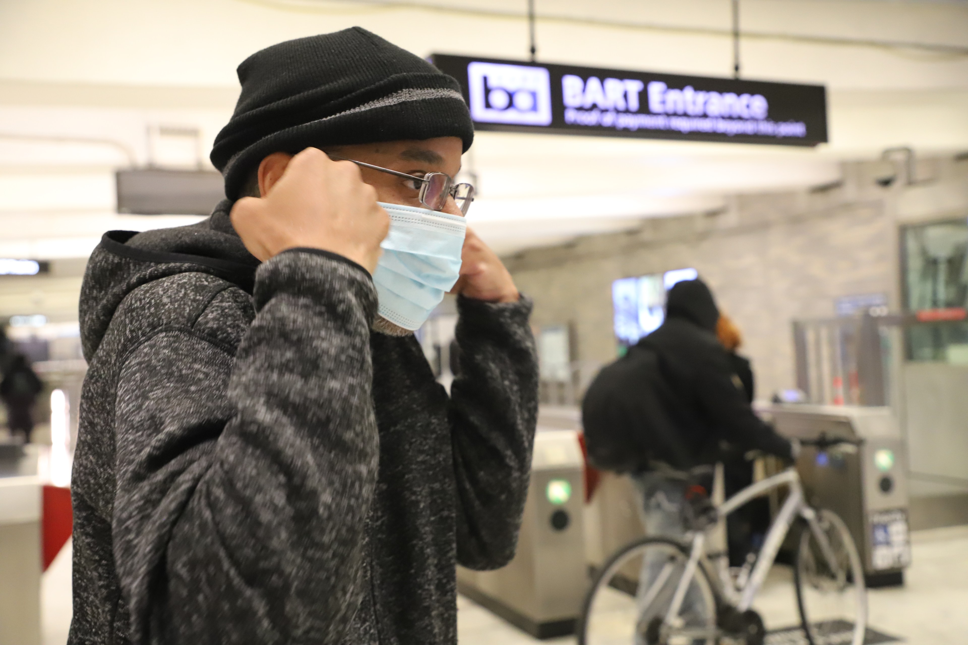 A person adjusts their mask in a station near a &quot;BART Entrance&quot; sign. Another individual with a bicycle is in the background.