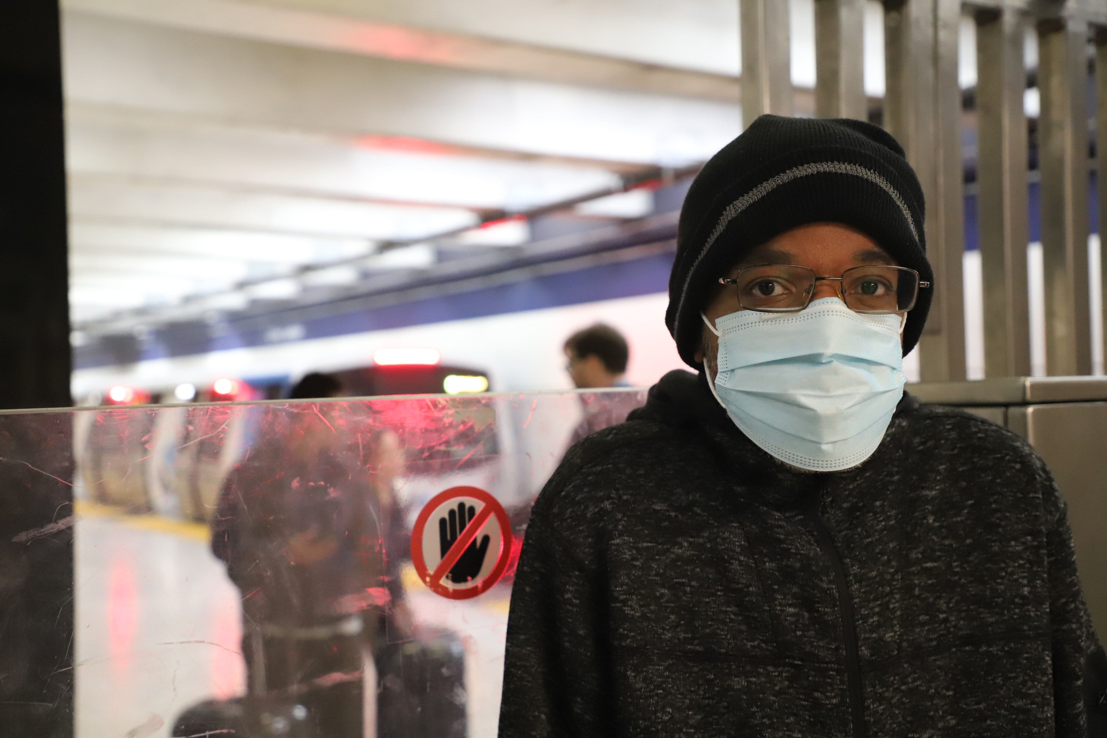 A person in a mask and beanie stands in a subway station with a blurry train in the background.