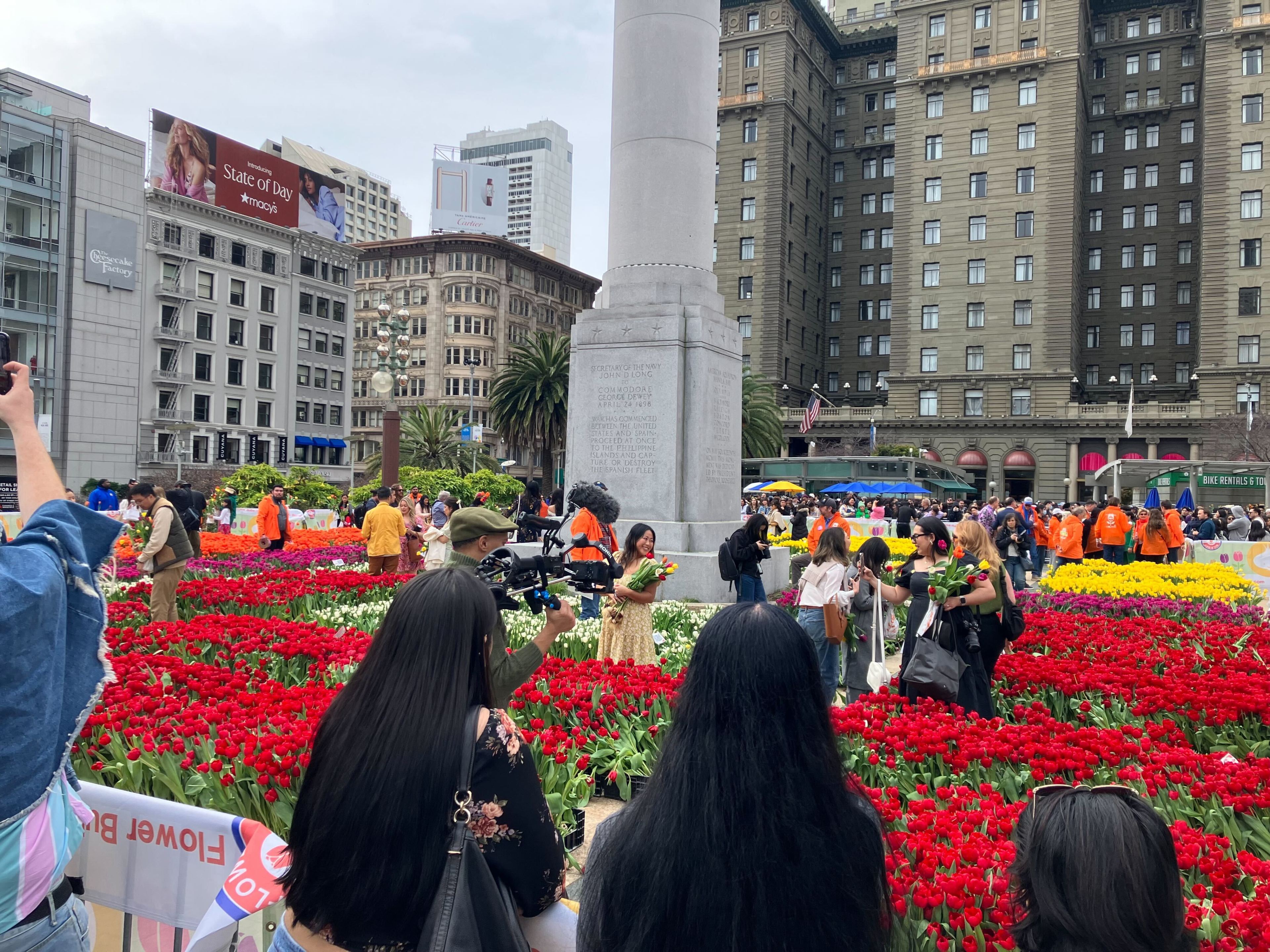 A bustling urban park with vibrant tulips and a crowd of people around a central monument, flanked by tall buildings.