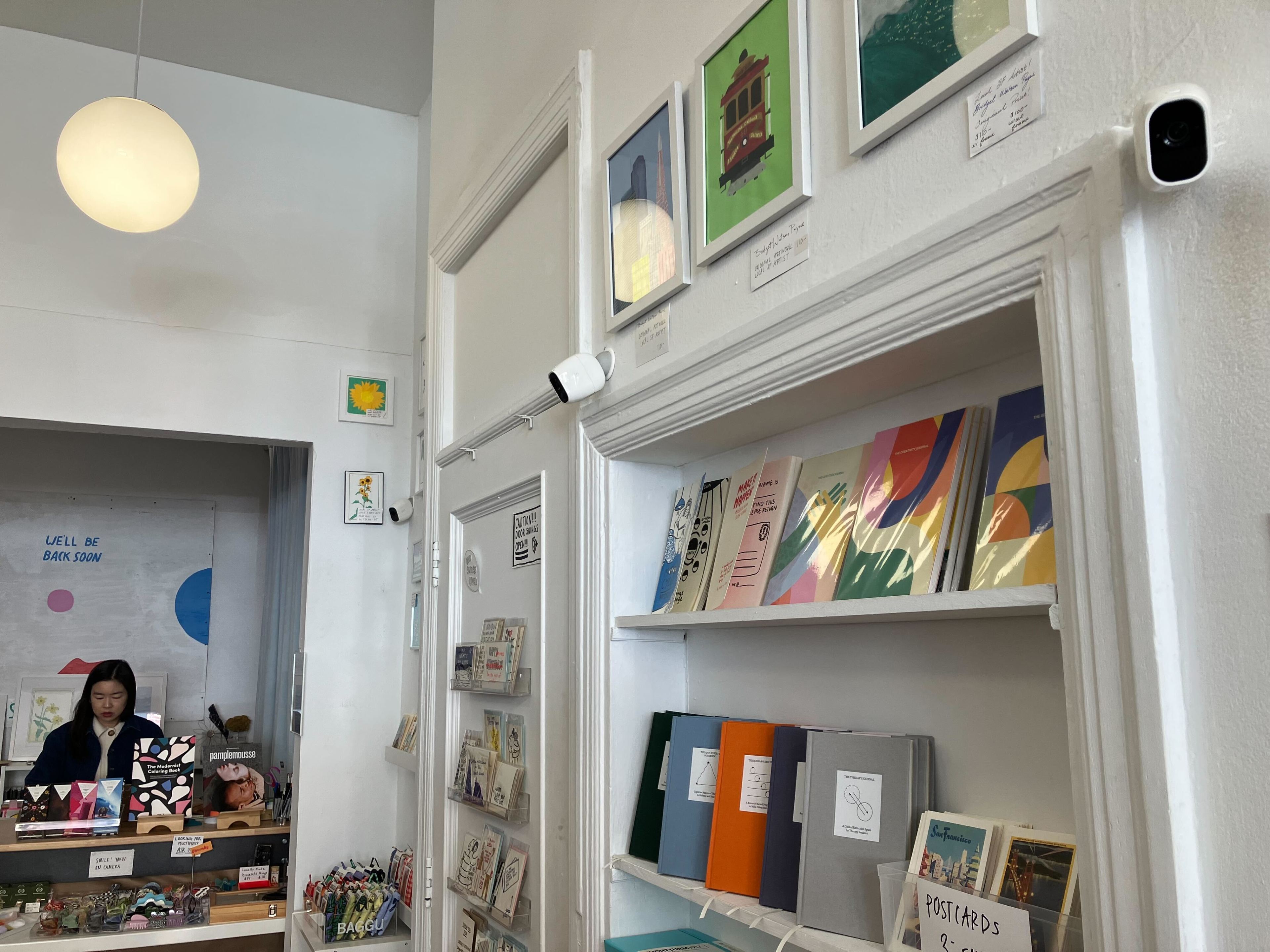 An art shop interior with prints, greeting cards on shelves, a white pendant light, and an attendant behind the counter.