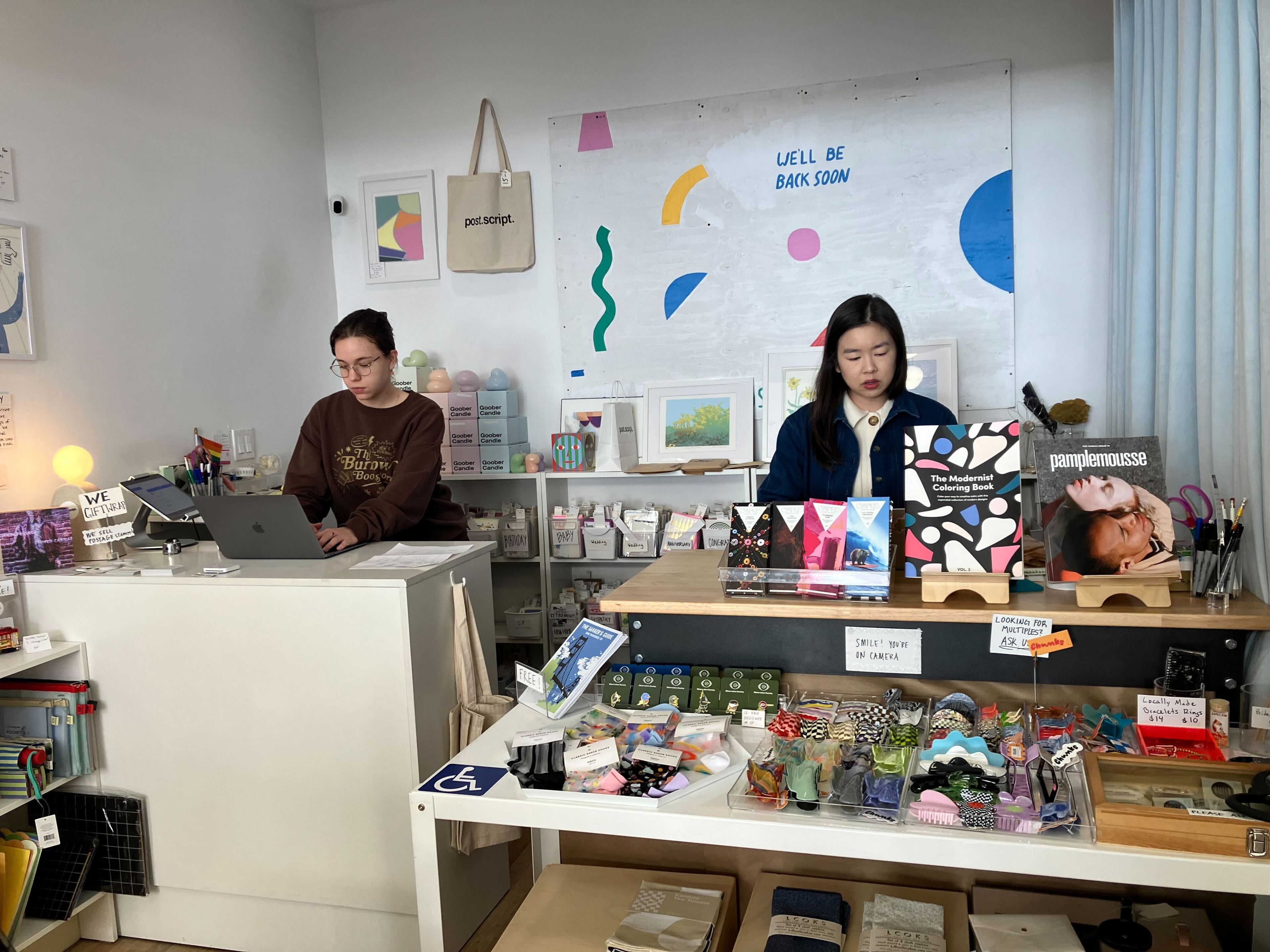 Two people work behind a counter in a bright, art-filled retail shop with various small items on display.