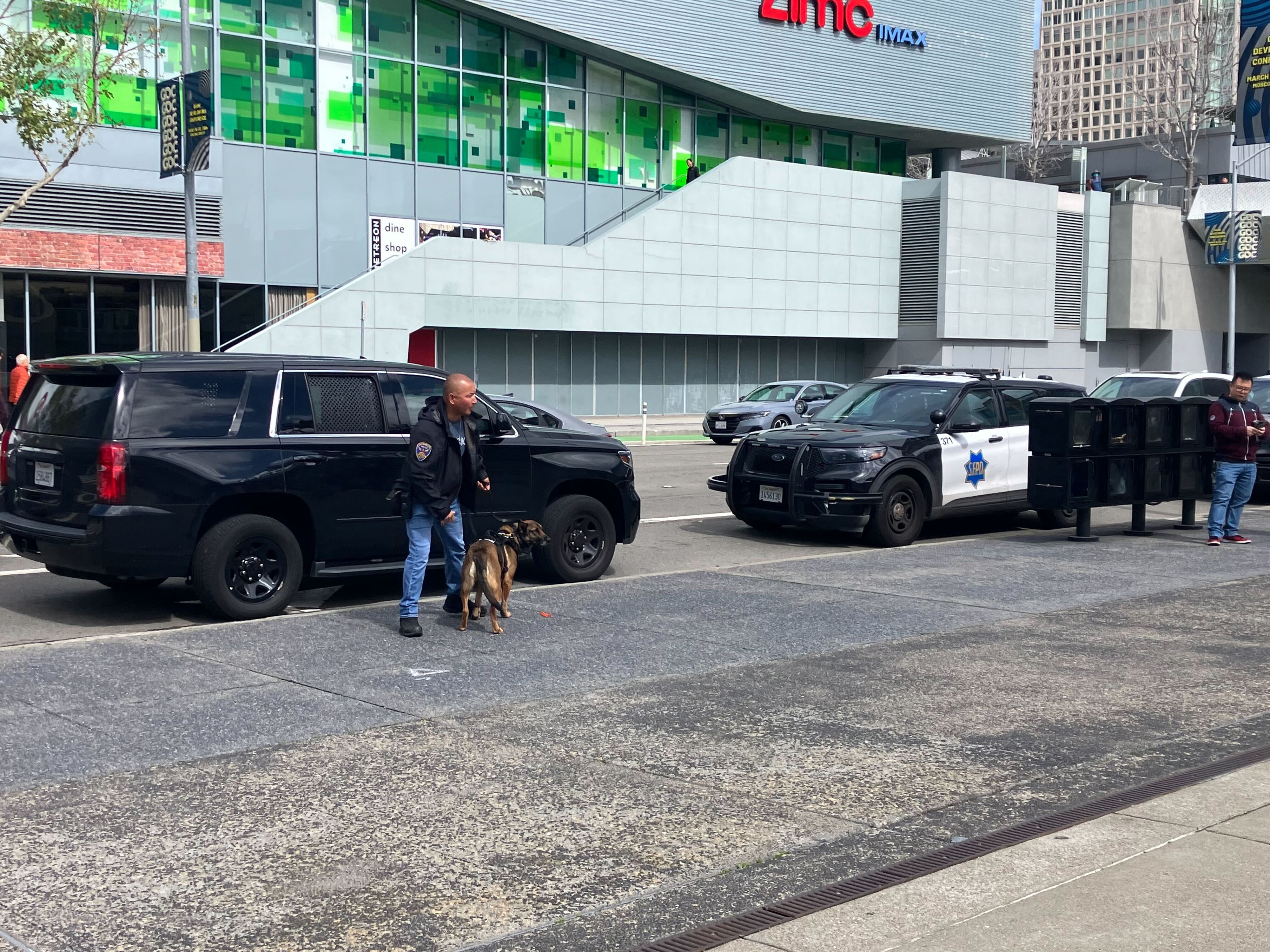 A security officer with a dog is walking beside a black SUV, with a police car and modern buildings in the background.