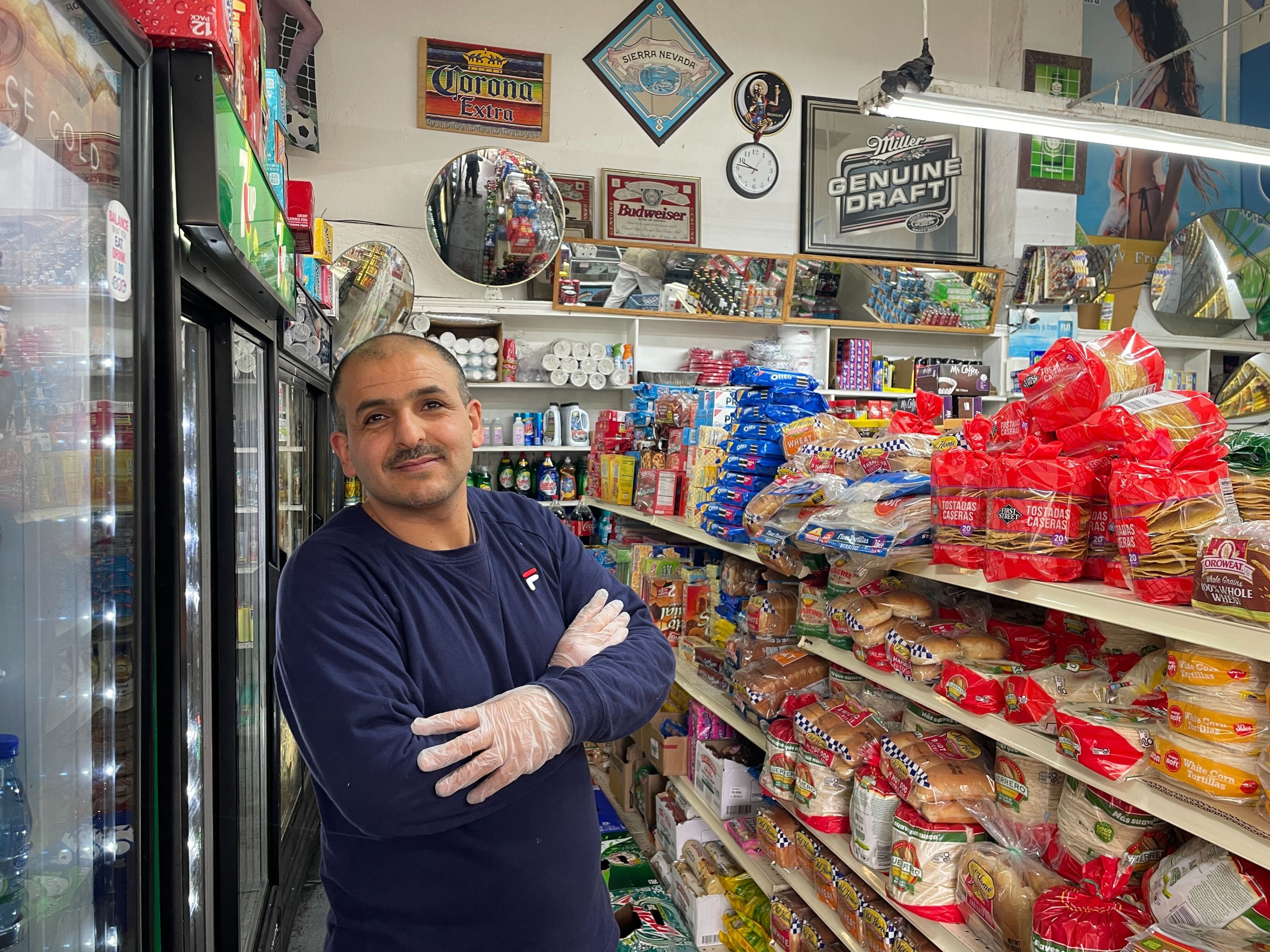 A man stands smiling in a store aisle, surrounded by snacks and beverages, wearing gloves and casual attire.