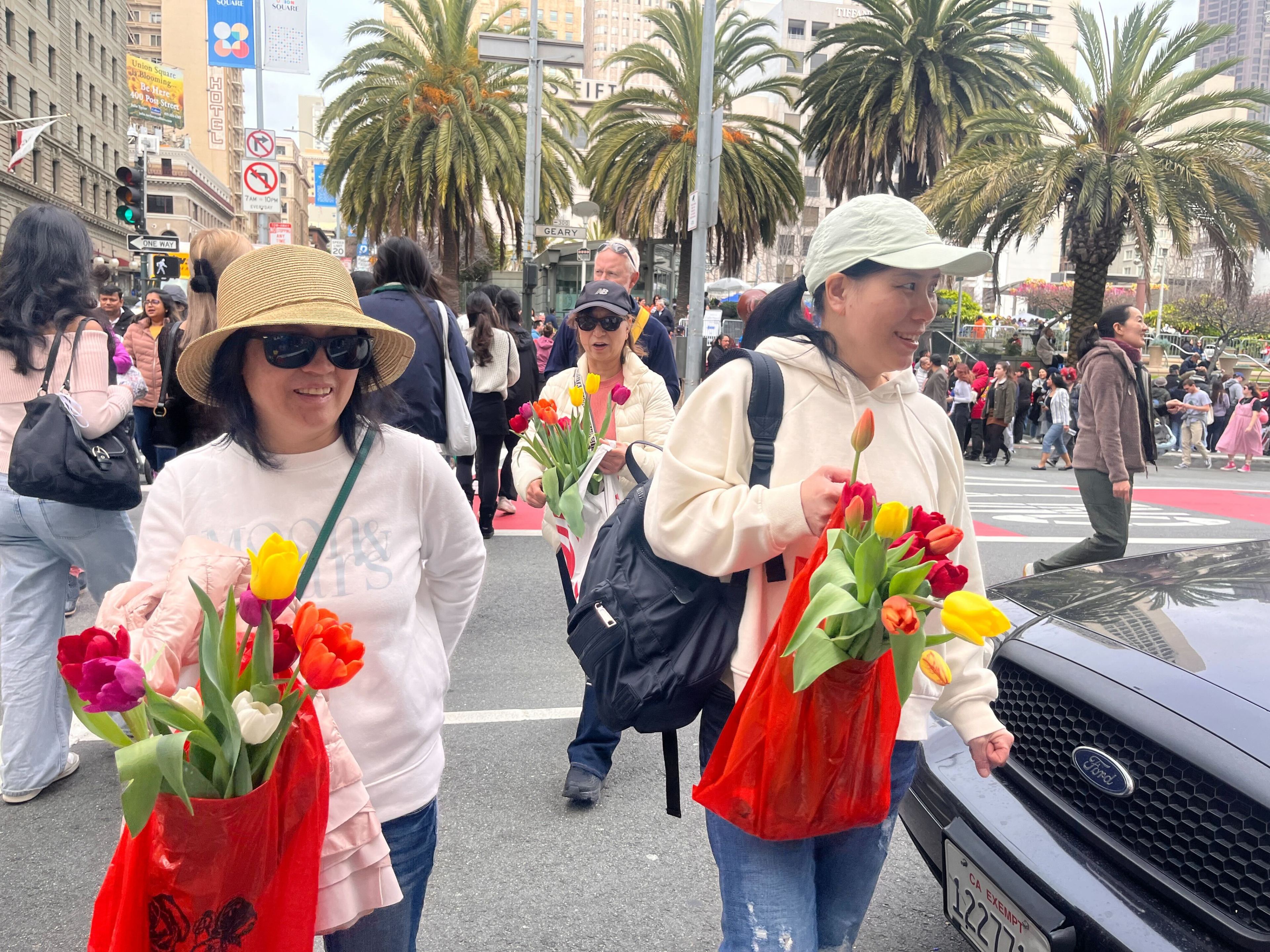 Two smiling women with hats and bouquets of tulips cross a busy city street with palm trees in the background.