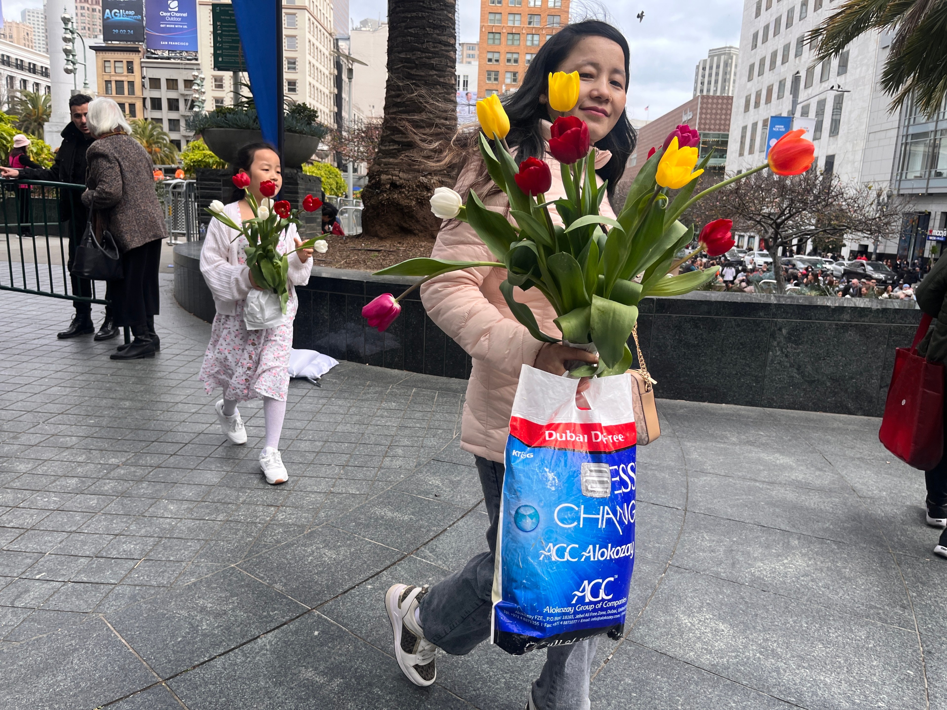 A woman holding colorful tulips in a plastic bag walks by a girl, both smiling, in an urban square.