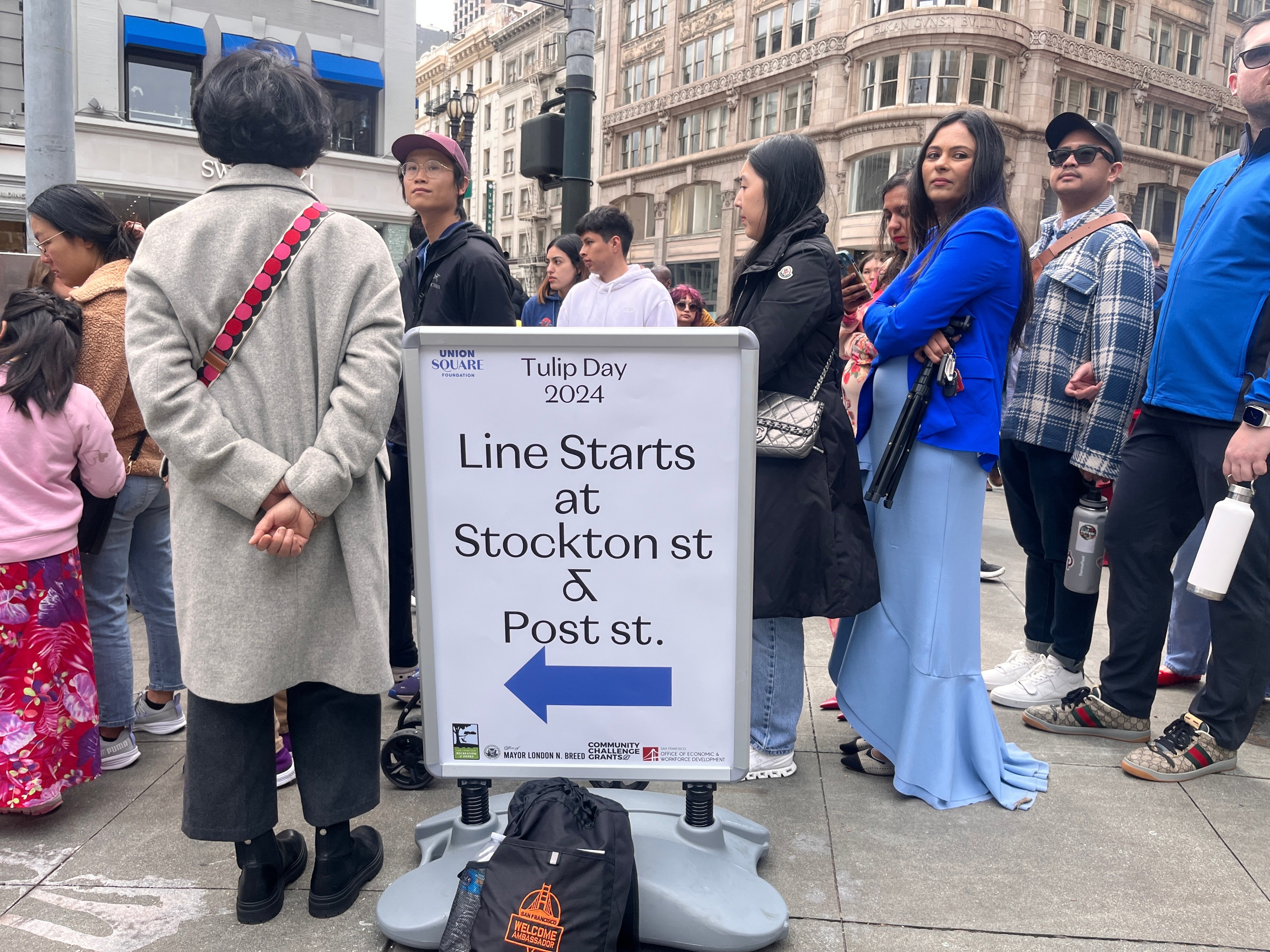 A diverse crowd stands by a sign indicating the start of a line for Tulip Day 2024 at an urban intersection.