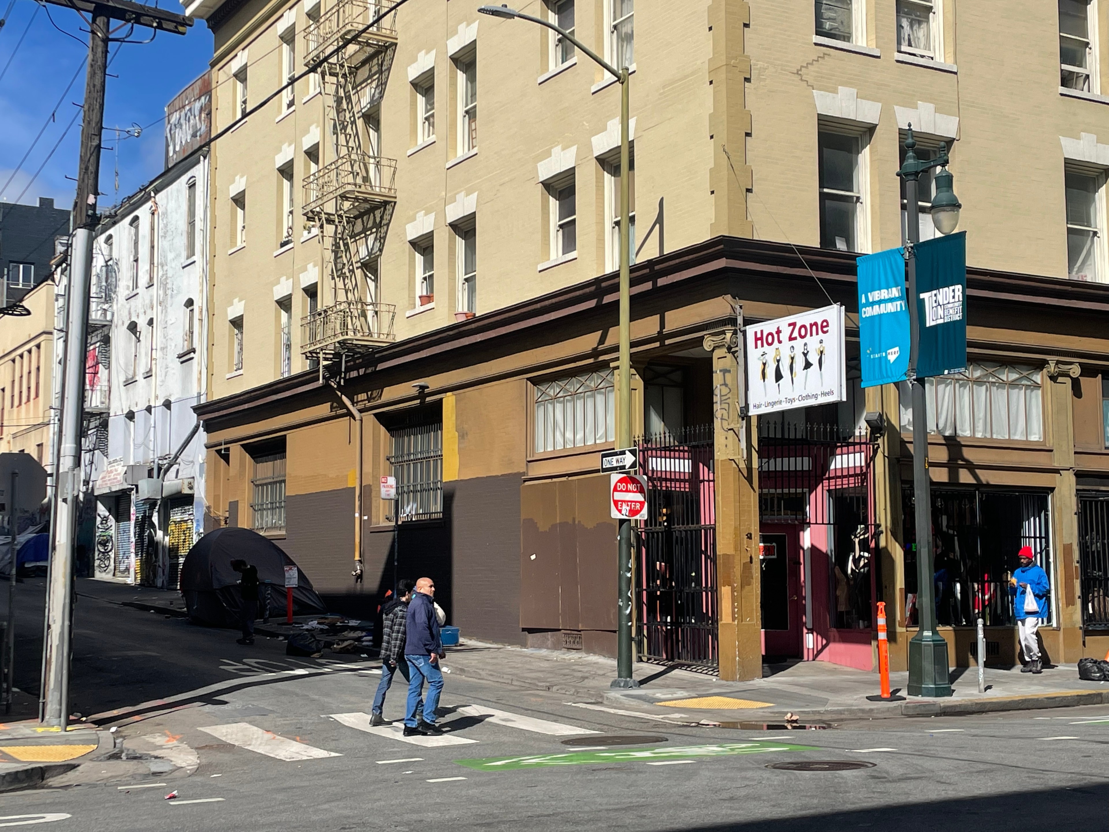 A sunny street corner with people, a yellow building, fire escapes, banners saying "Hot Zone" and "Vibrant Community," plus a film crew tent.
