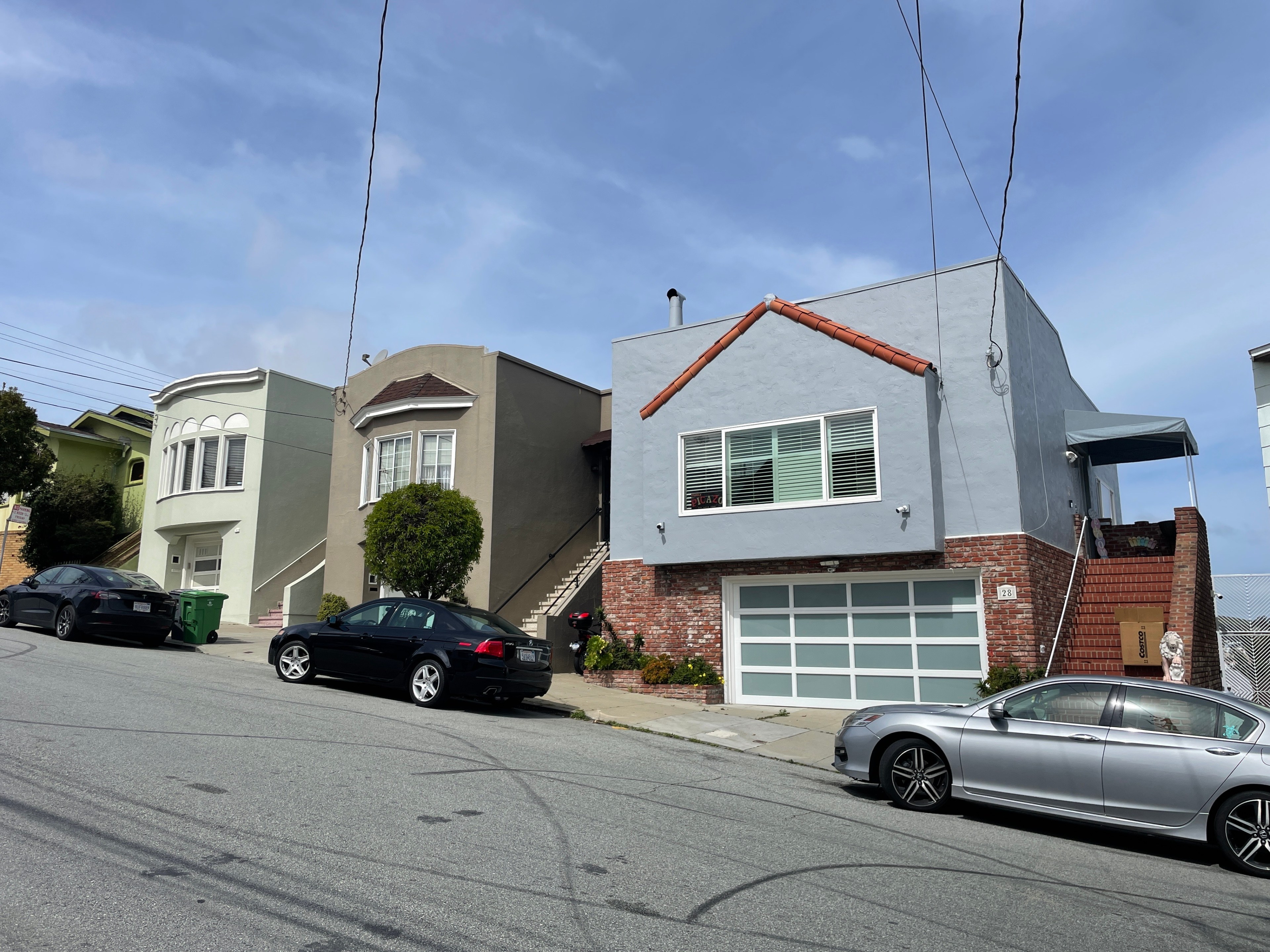 Three houses on a steep hill with parked cars, under a partly cloudy sky.