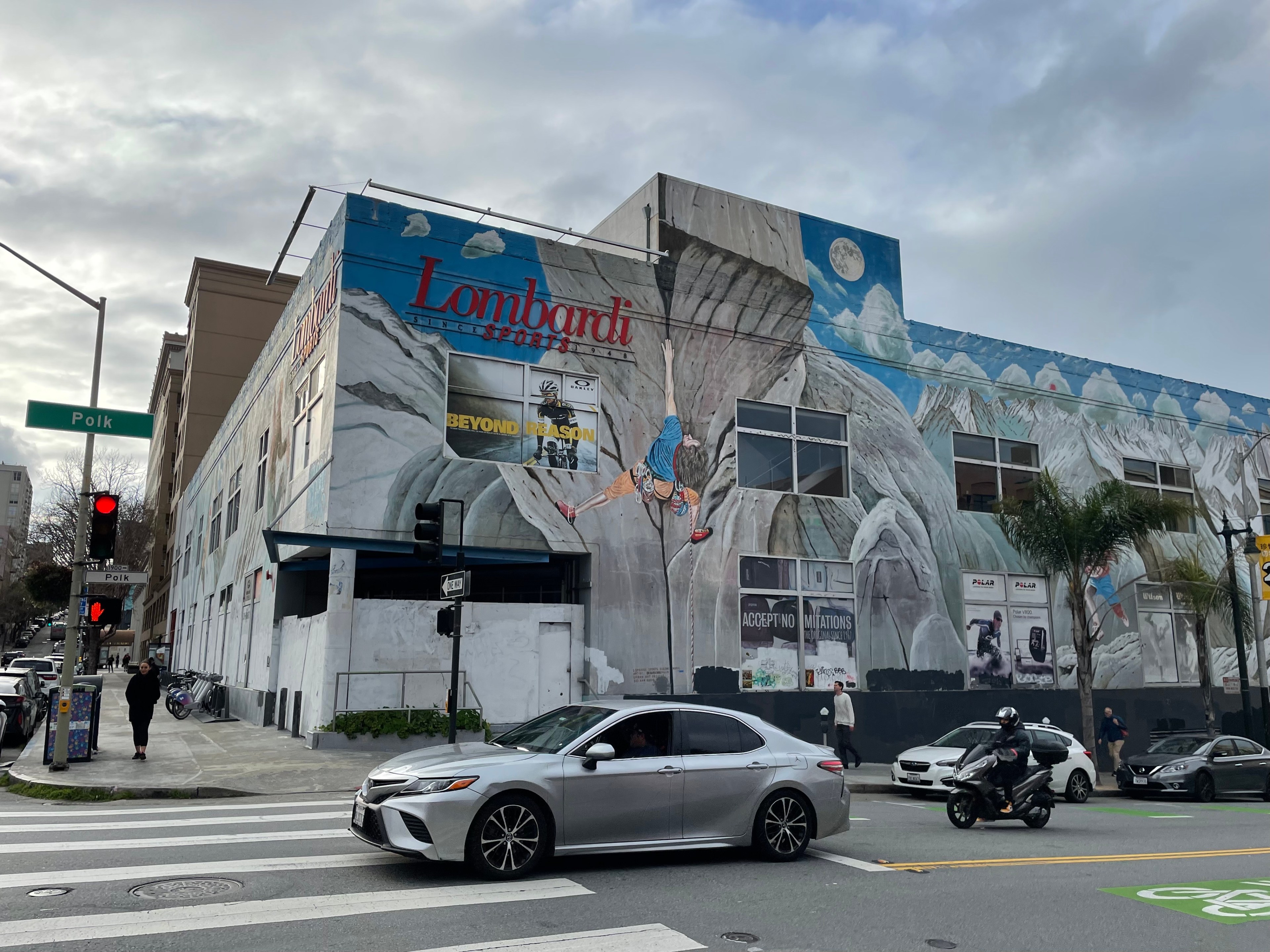 Cars pass in front of a large, empty retail building covered in a mural depicting winter sports.