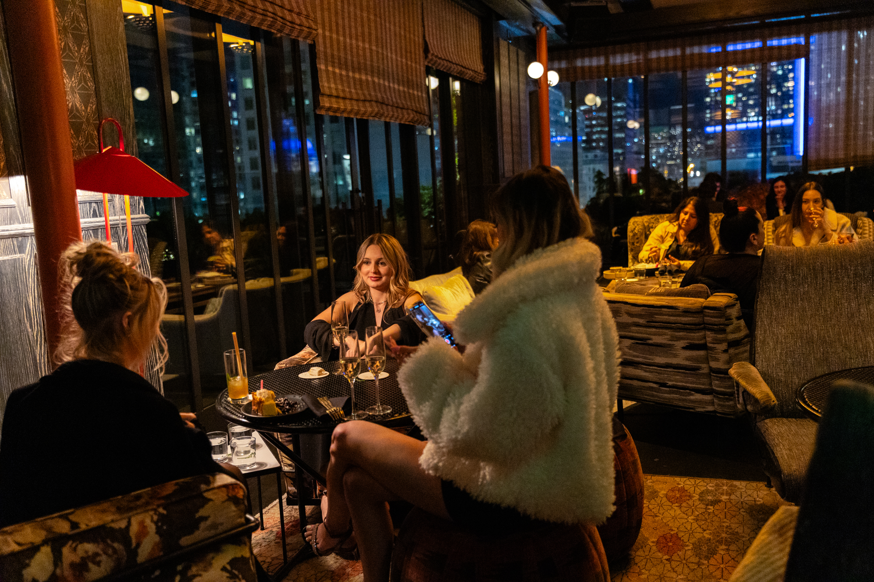 People socialize in a cozy night-time lounge with a cityscape backdrop. Drinks on tables, warm lighting, and a relaxed ambiance.