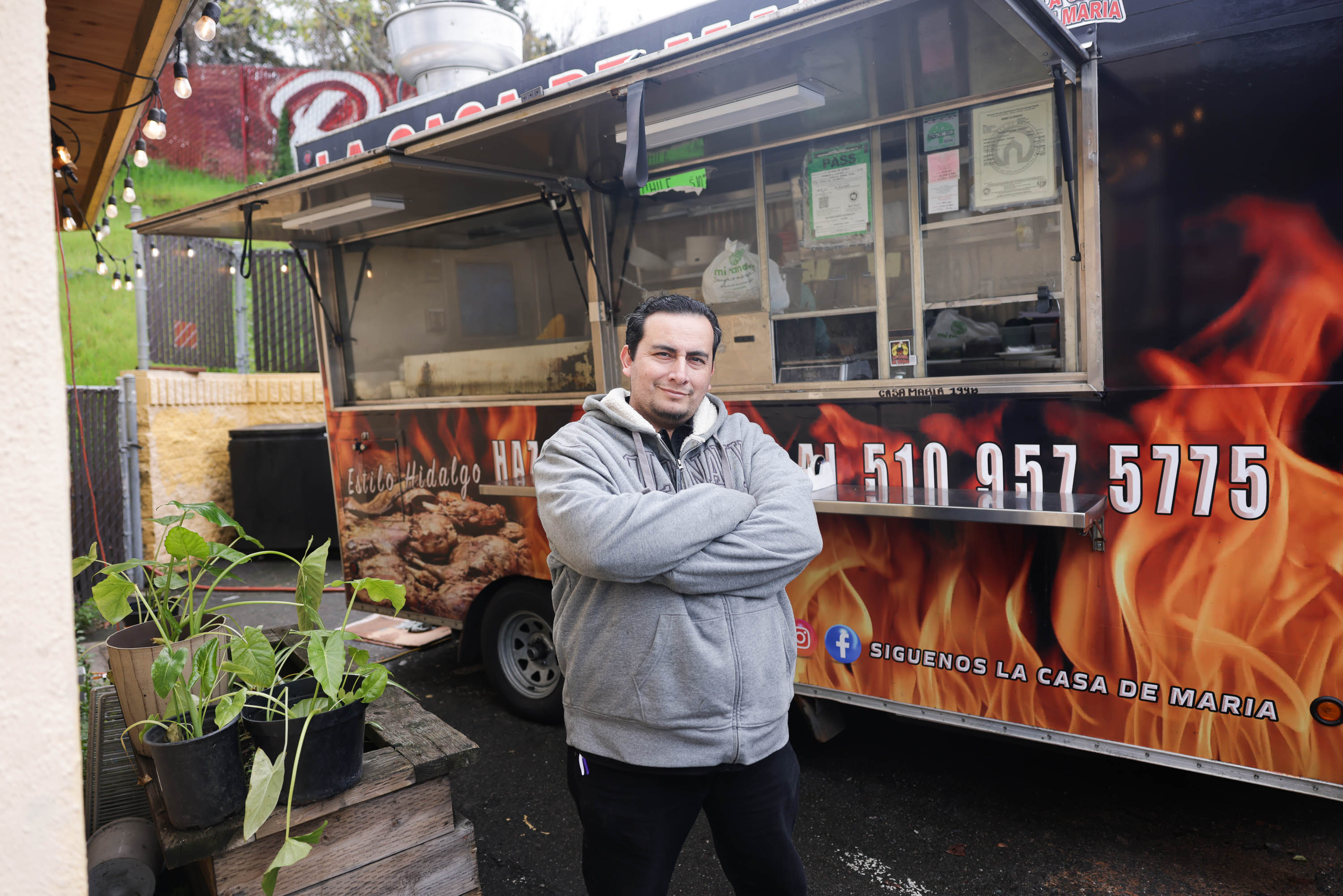 Man with arms crossed stands in front of a food truck with fiery graphics.