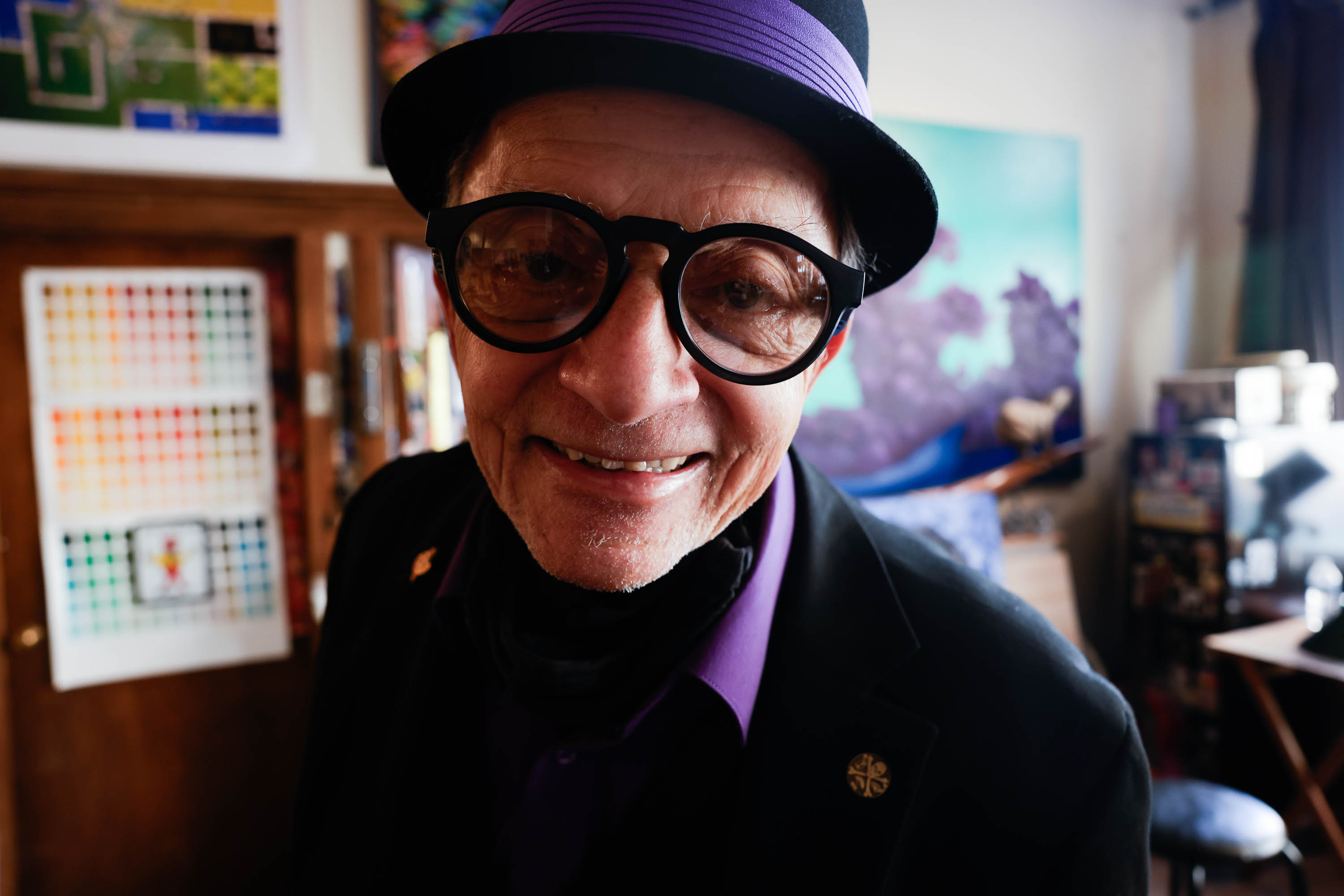 An elderly man in large black glasses and a purple hat smiles closely at the camera, with art and a cozy room in the background.