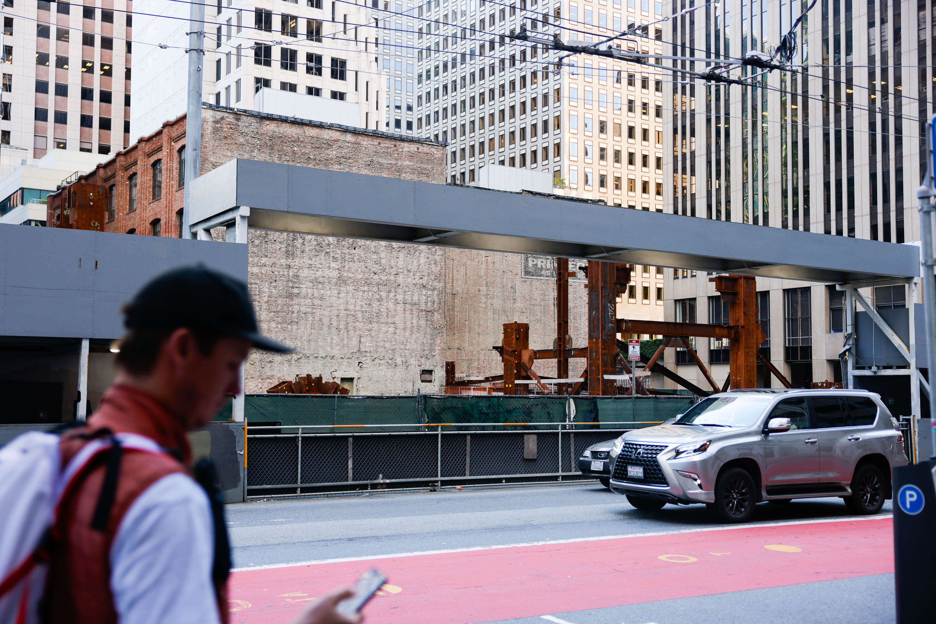 An urban construction site with exposed steel beams, surrounded by a mix of old and modern buildings, a walking man in foreground, and cars on the street.