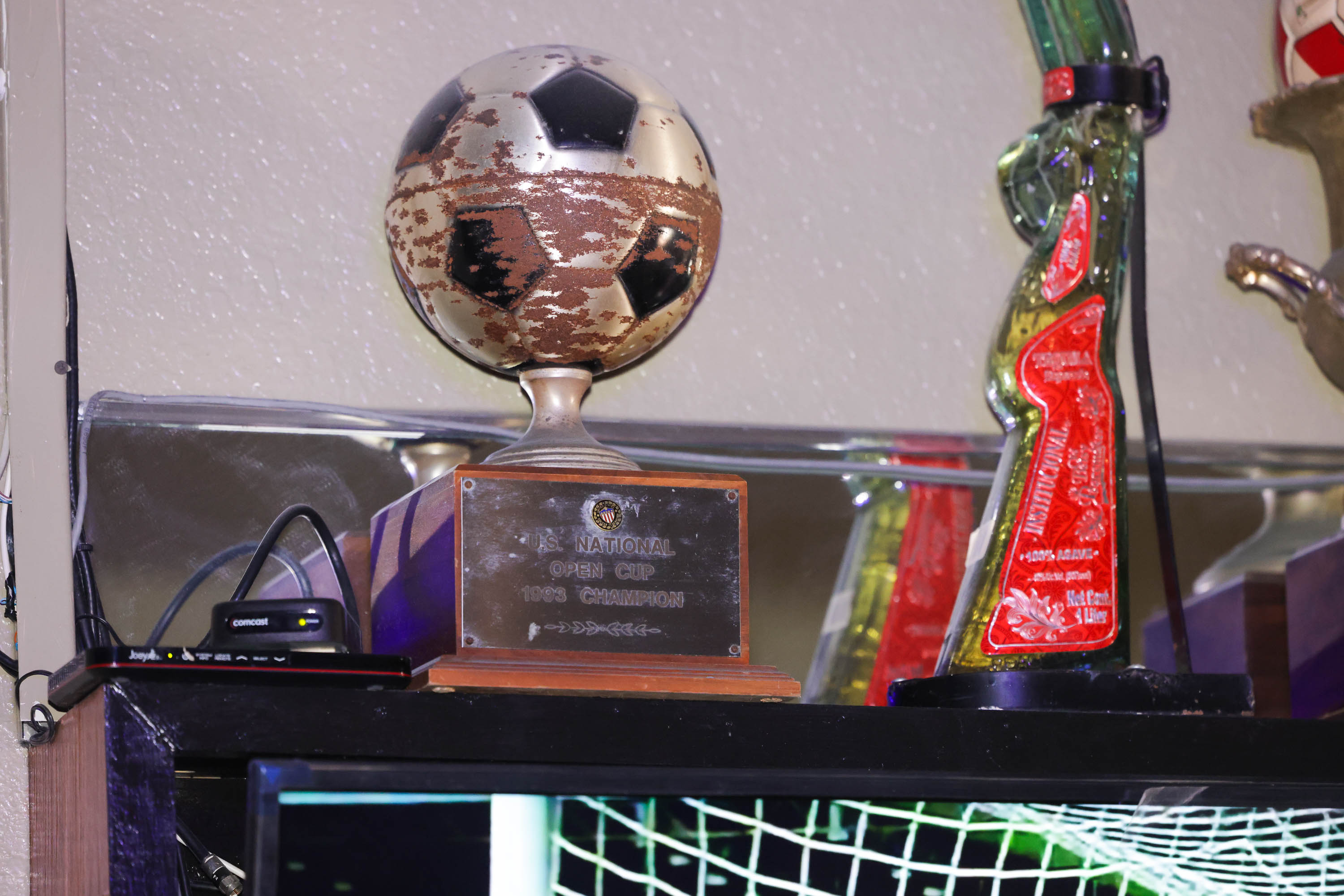 A shelf holds a weathered soccer ball trophy labeled &quot;1993 Champion&quot; and a colorful glass award next to electronic devices.