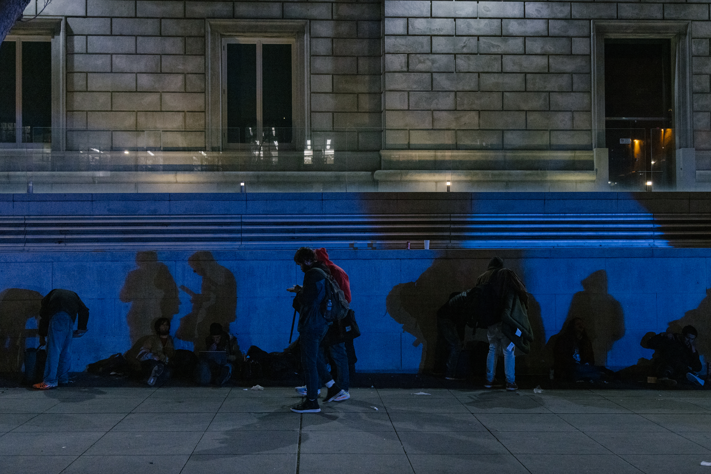 People casting shadows on a blue-lit building wall at night; some are seated, one stands using a phone.