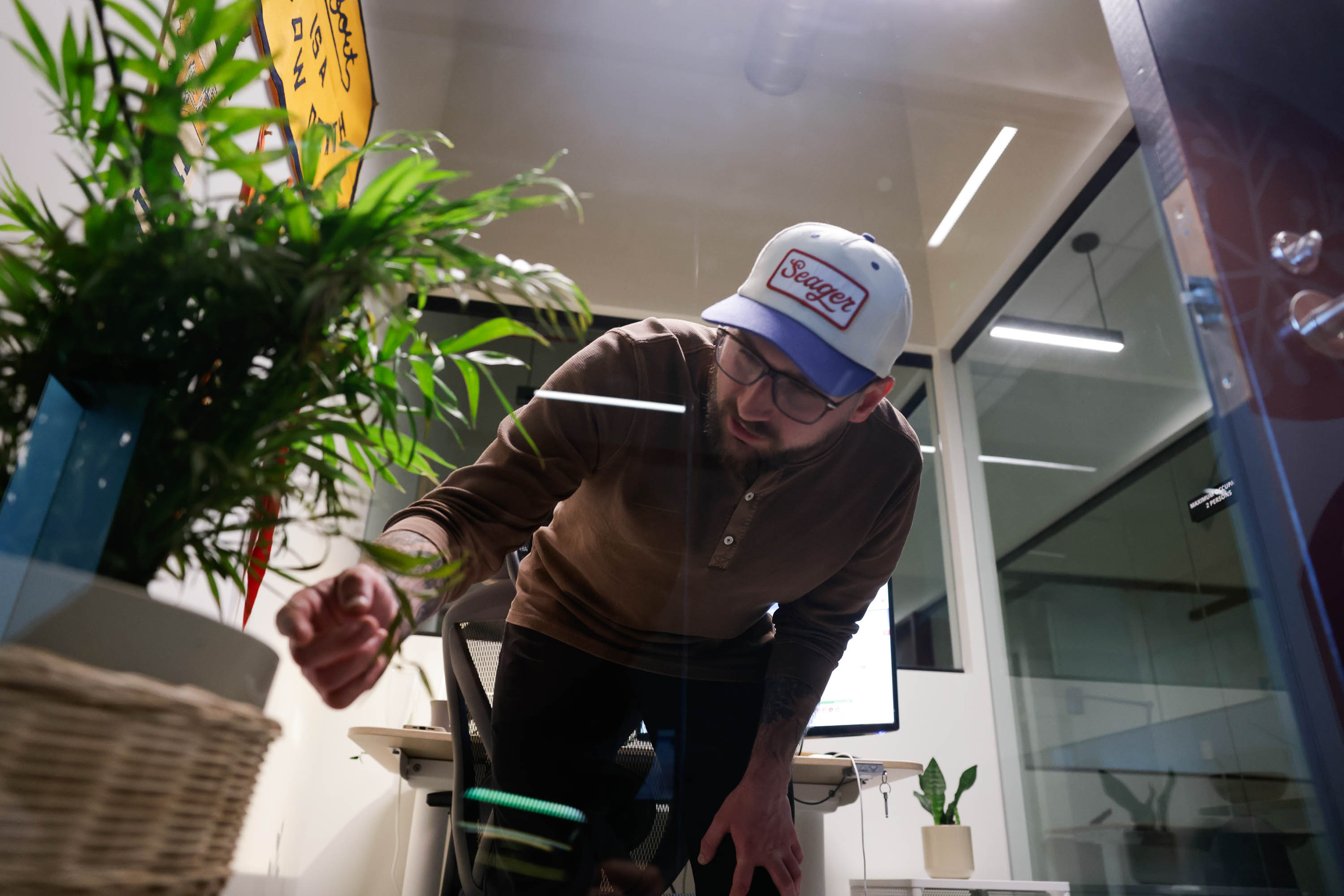 A man in a cap leans over a chair in an office, with plants and a glass door nearby.
