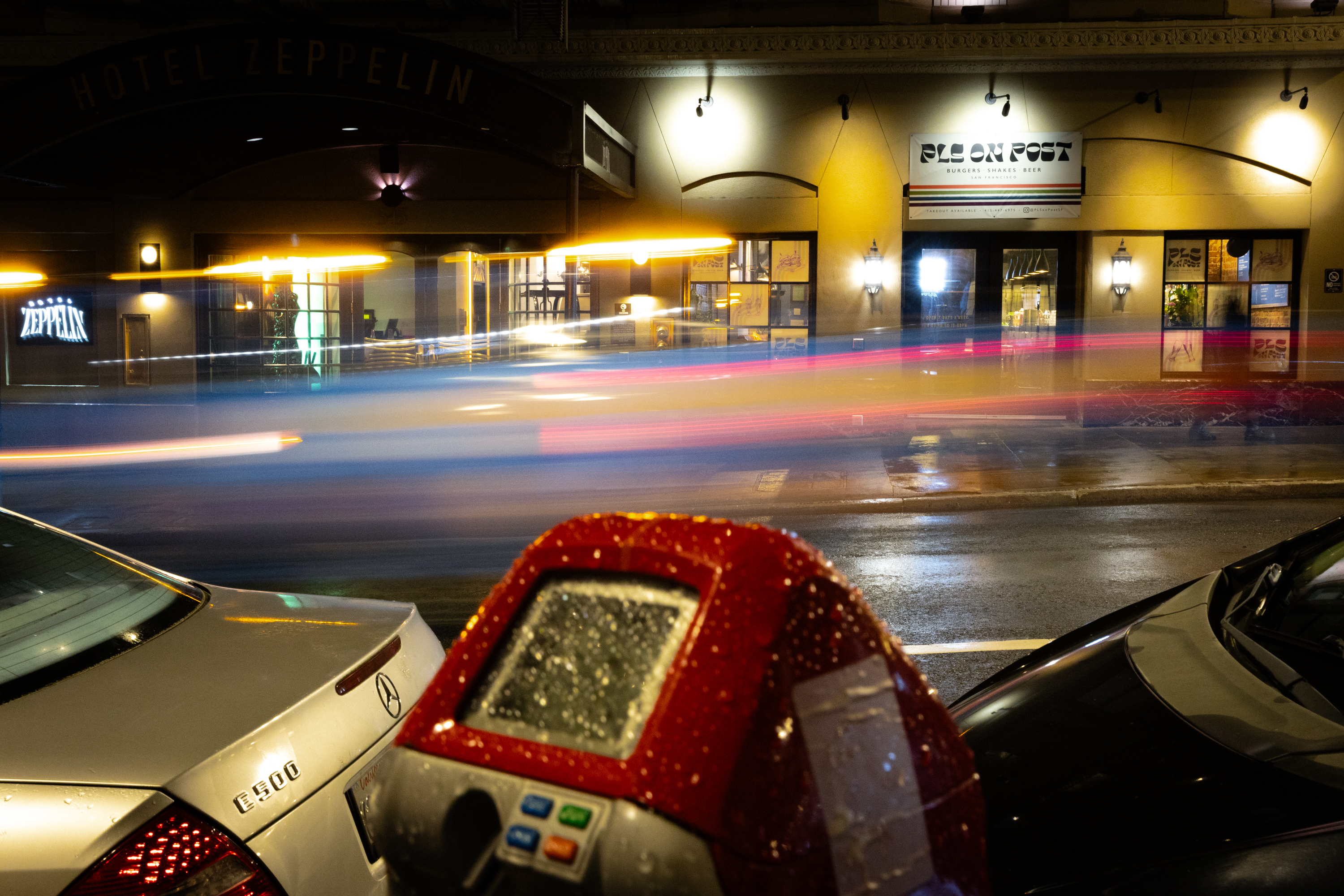 A nighttime street scene with a light trail blur from a moving vehicle, a wet parking meter, and parked cars in front of a building.