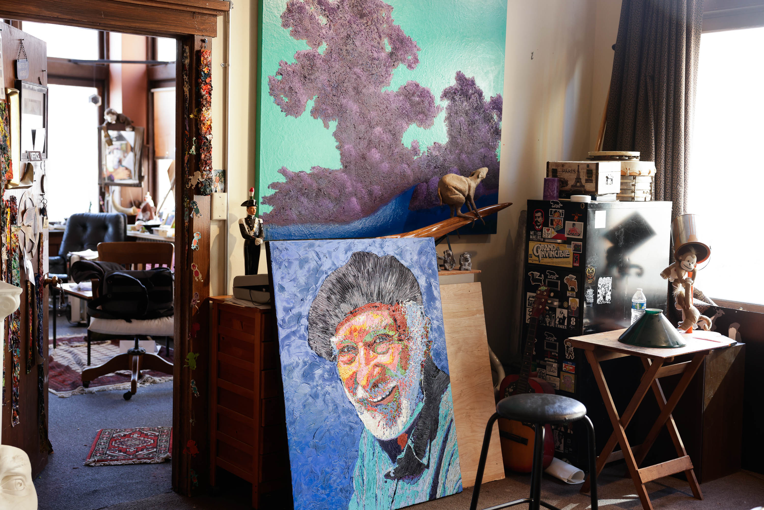 An artist's studio with colorful paintings, eclectic decor, and a portrait of a smiling man on an easel.