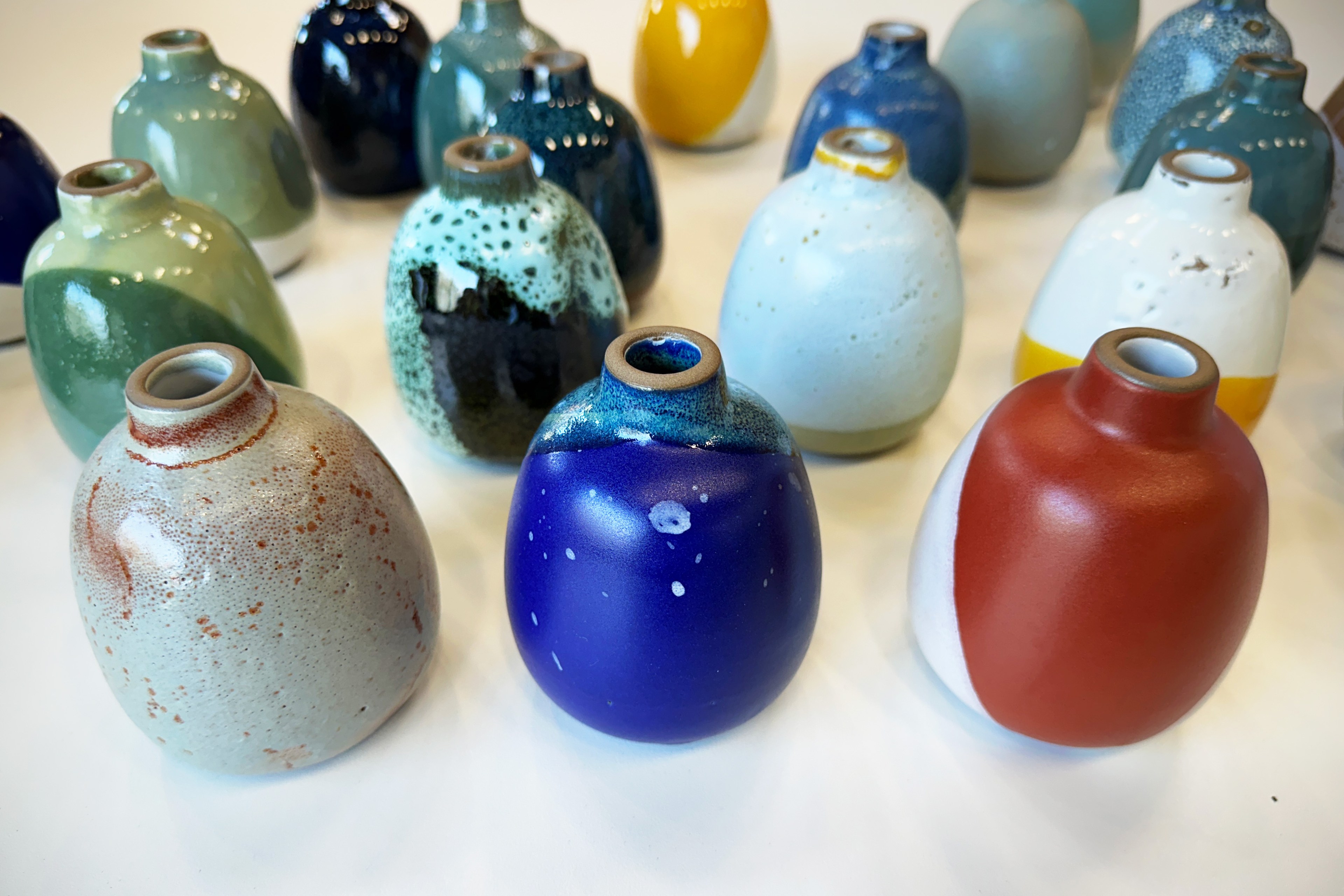 A collection of colorful ceramic vases of various colors sit on a white surface.